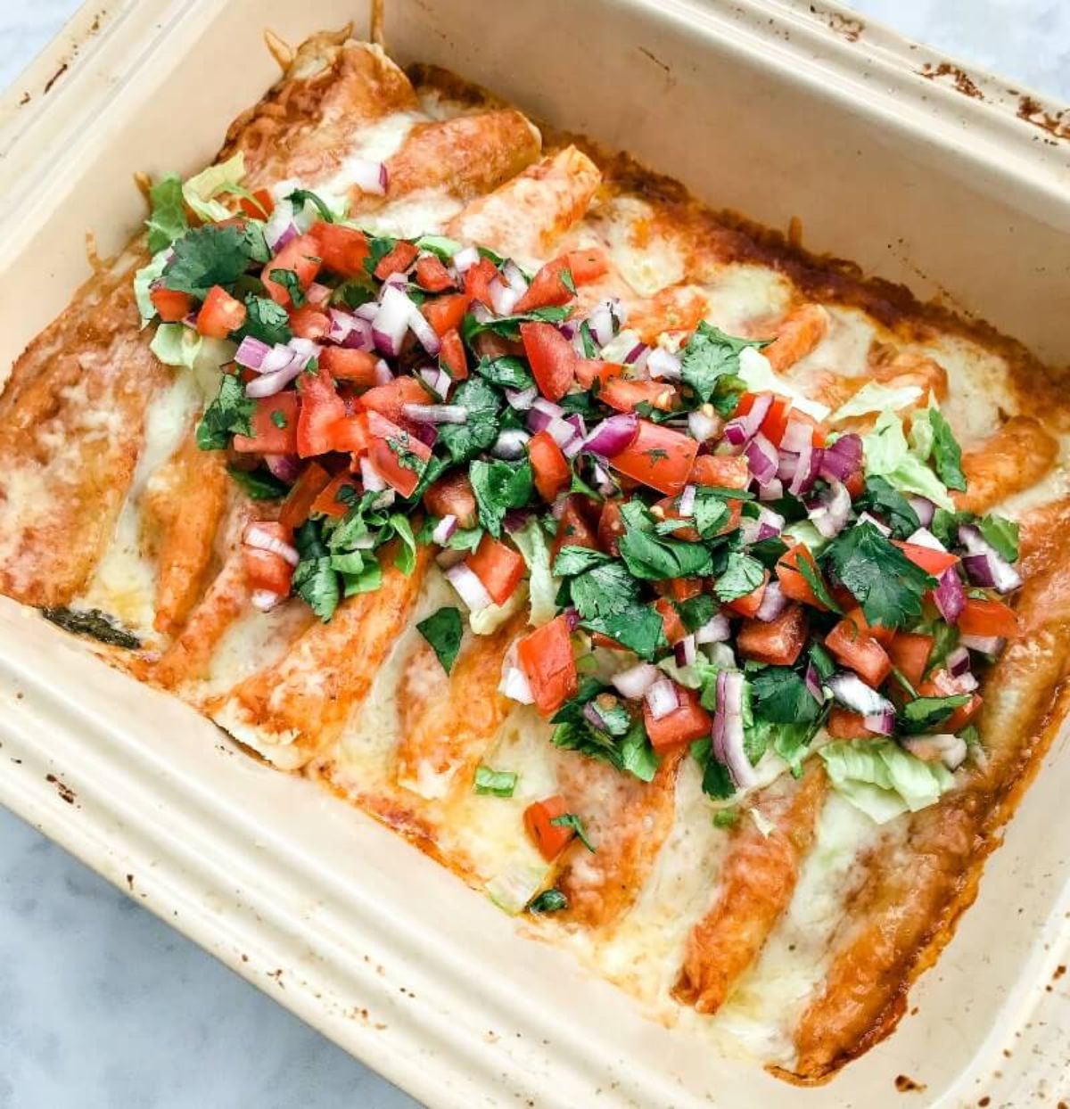 An oven dish filled with enchiladas topped with chopped fresh tomatoes, red onion, cucumber and herbs