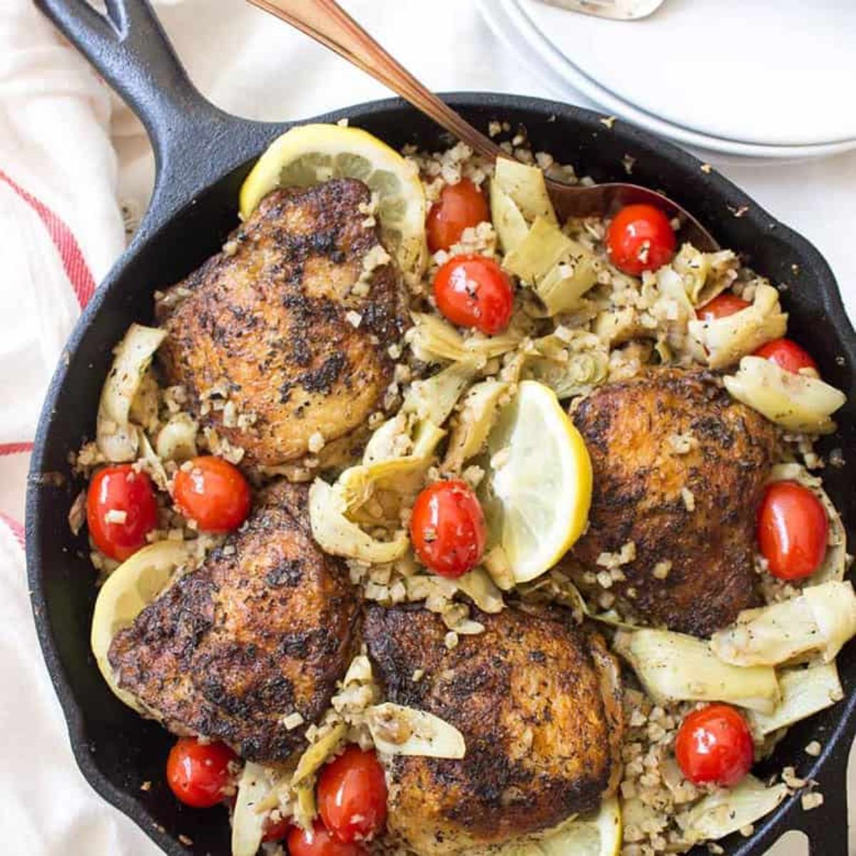 a skillet full of chicken thighs on a bed of vegetables, lemon slices and plum tomatoes