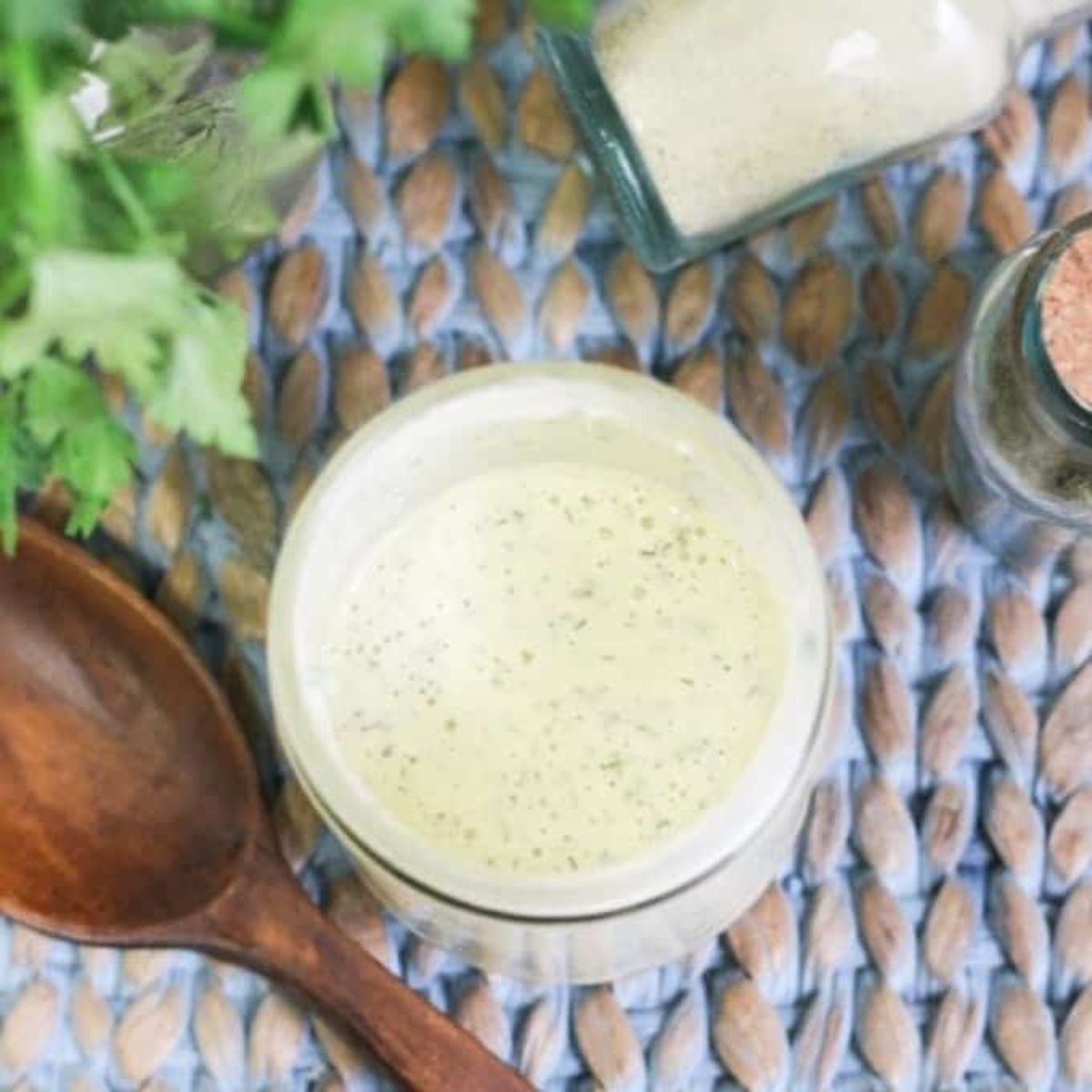 a jar of jalapeno ranch dressing on a raffia mat with a wooden spoon and some herbs next to it