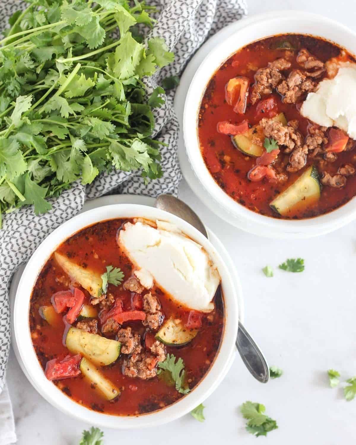 2 bowls of lasagne soup topped with mozzarella slices. A bunch of cilantro is to one side