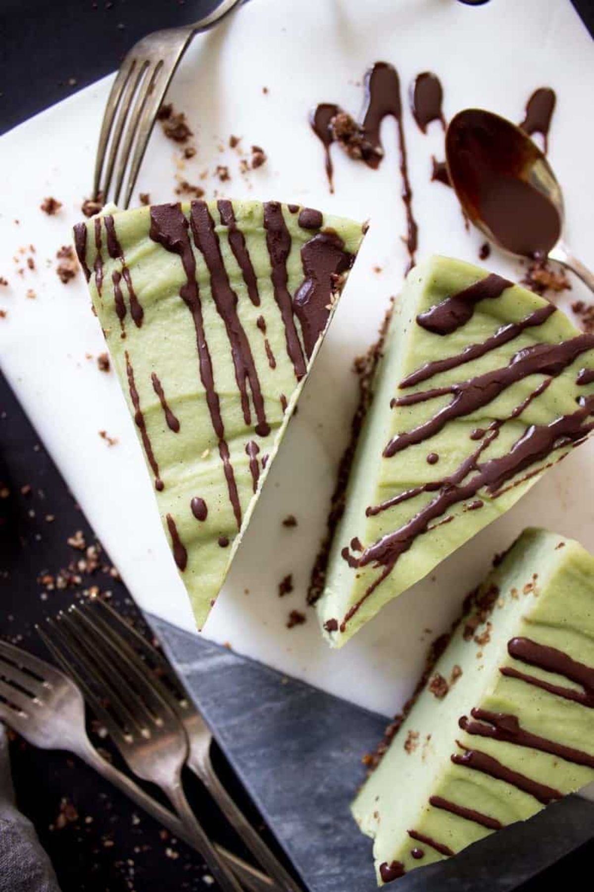 A tile with 3 slices of light green colored cheesecake slices drizzled with chocolate sauce