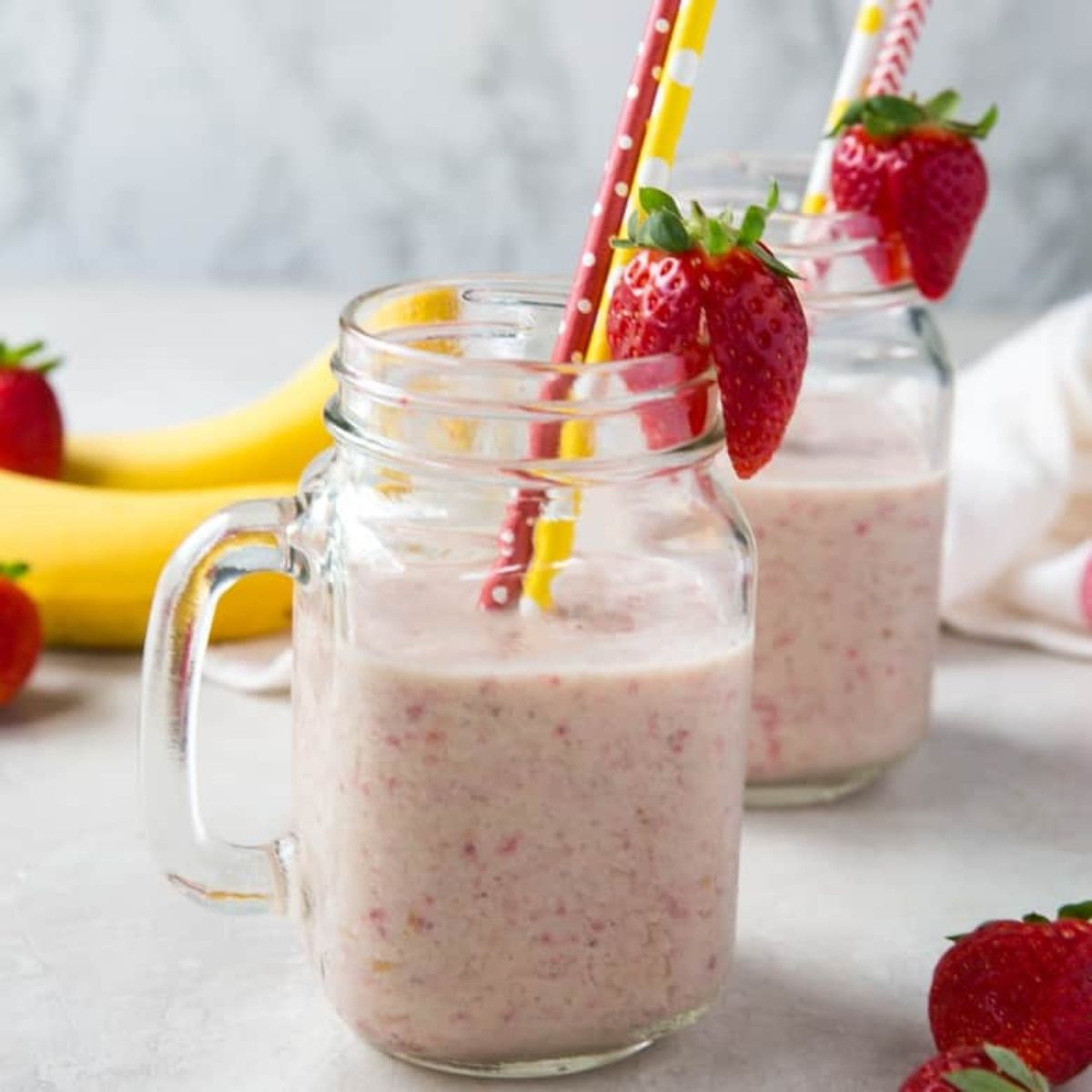 2 squared mason jars with strawberry banana smoothie in them, garnished with whole strawberries and each with a red and yellow straw in them