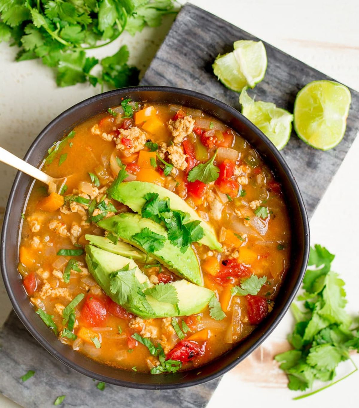 a bowl of turkey taco soup topped with avocado slives and chopped cilantro. A silver spoon rests in the soup and lime wedges and herbs are scattered around