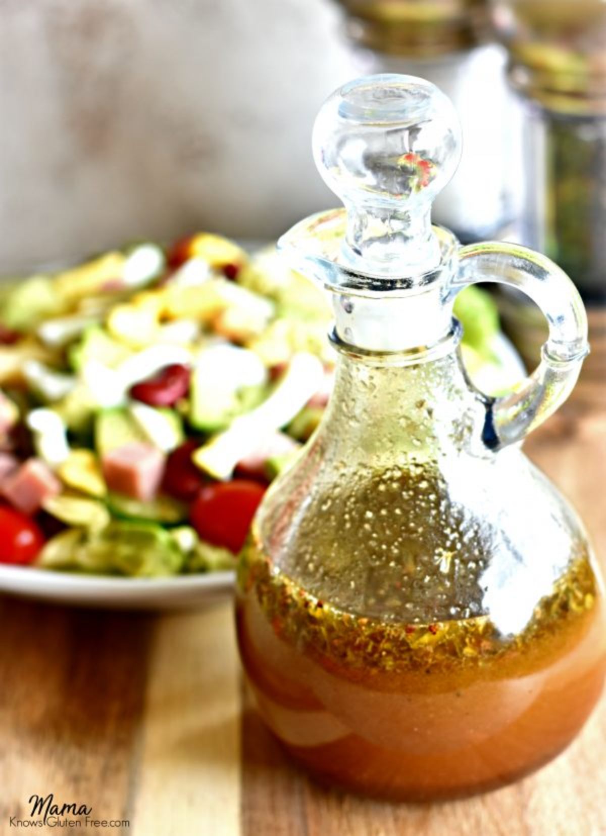 A glass jug with stopper full of balsamic vinaigrette with a plate of salad behind it
