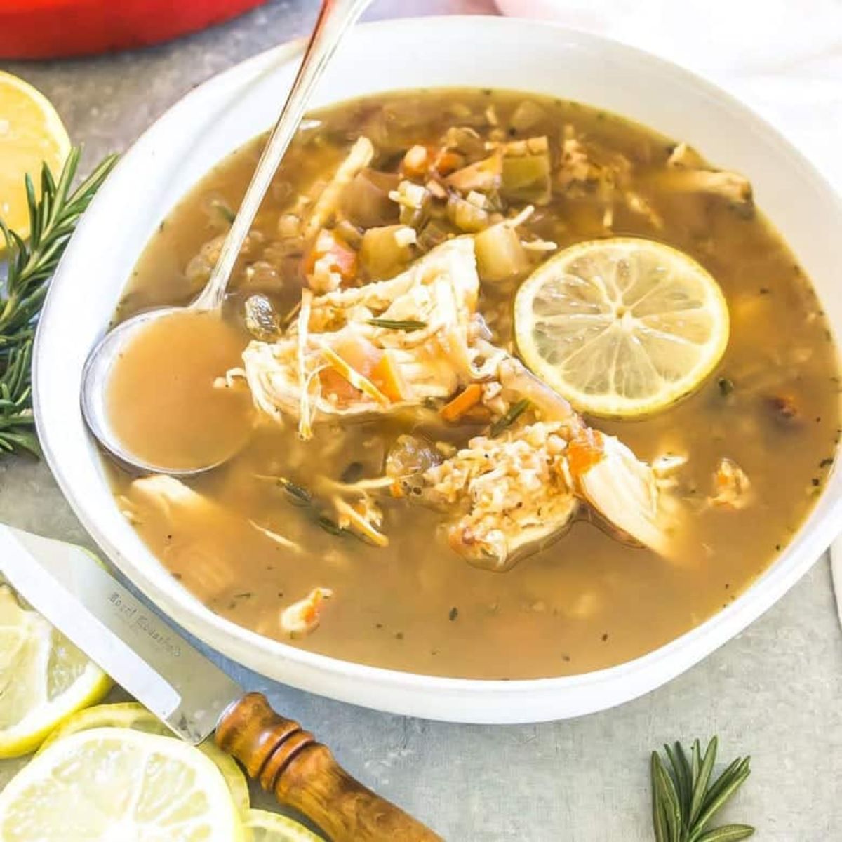 a white bowl of lemon chicken soup with lemon slices seen floating. A silver spoon is resting in the bowl and a wooden handled knife is below next to some more lemon slices