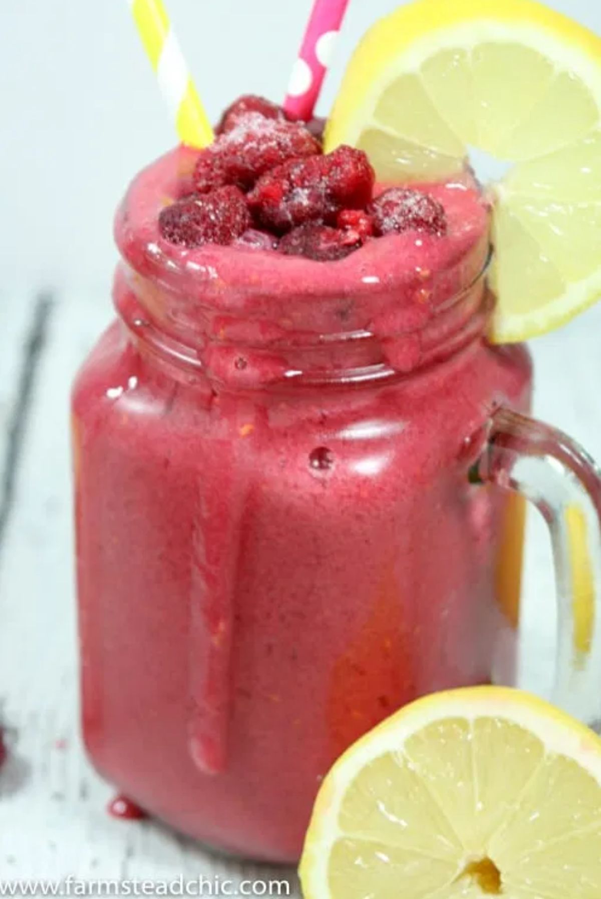 a square mason jar contains a raspberry lemonade smoothie, topped with frozen berries and garnished with a lemon slice