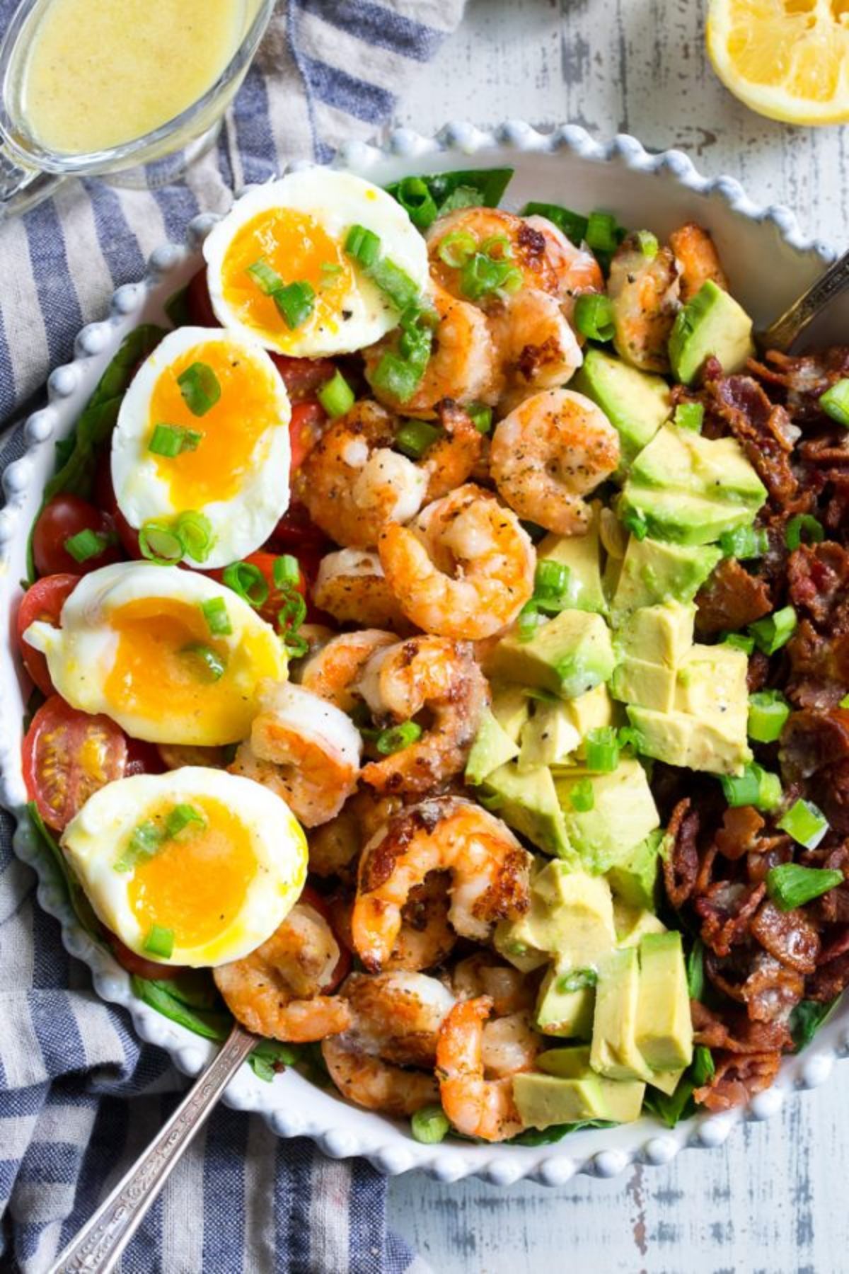 A white pie dish filled with shrimp, bacon, avocado, tomatoes and halved boiled eggs, sprinkled with scallions