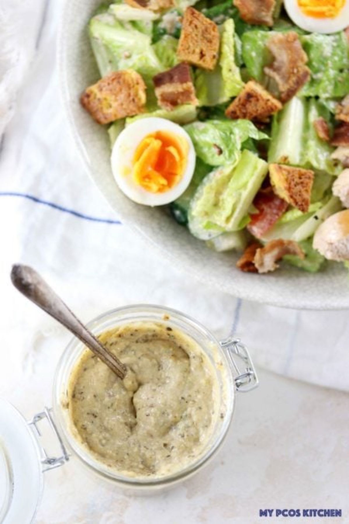 A glass jar of thick cream y dressing with a spoon in it, in front of a bowl of salad