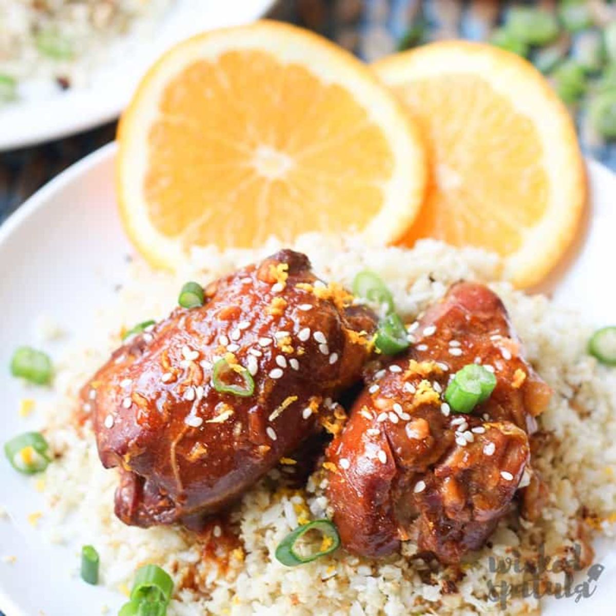 a plate of marinaded chicken thighs, on a bed of cauliflower rice garnished with sliced scallions, sesame seeds and orange slices