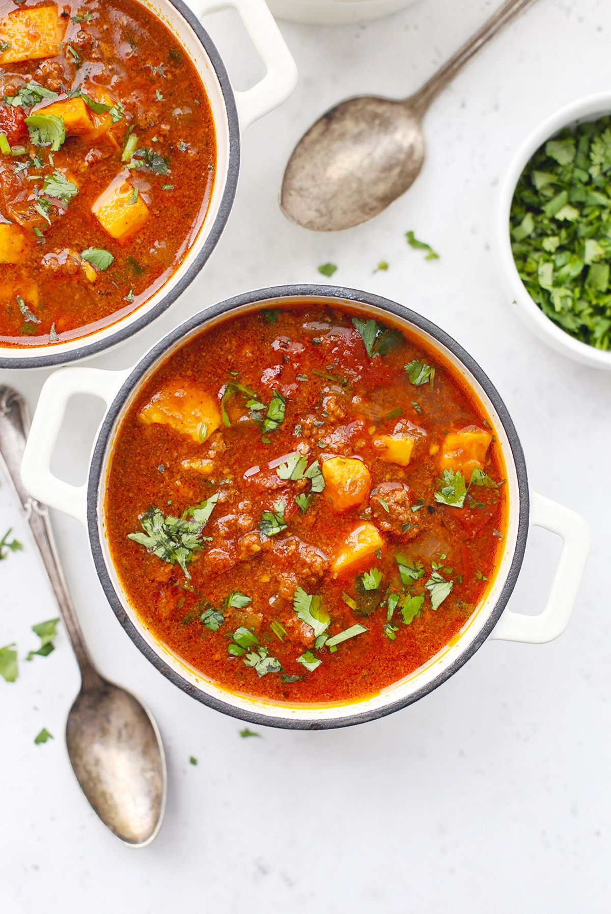 2 handled bowls filled with sweet potato chilli garnsihed with chopped herbs, and spoons next to them