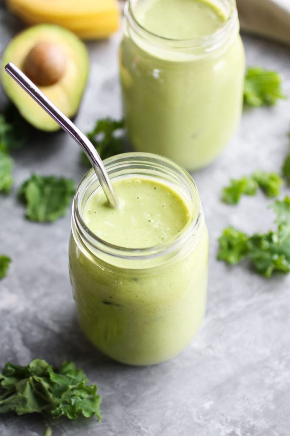 2 mason jars of green smoothie, one with a metal straw sticking out of it. Kale leaves and half an avocado can be seen