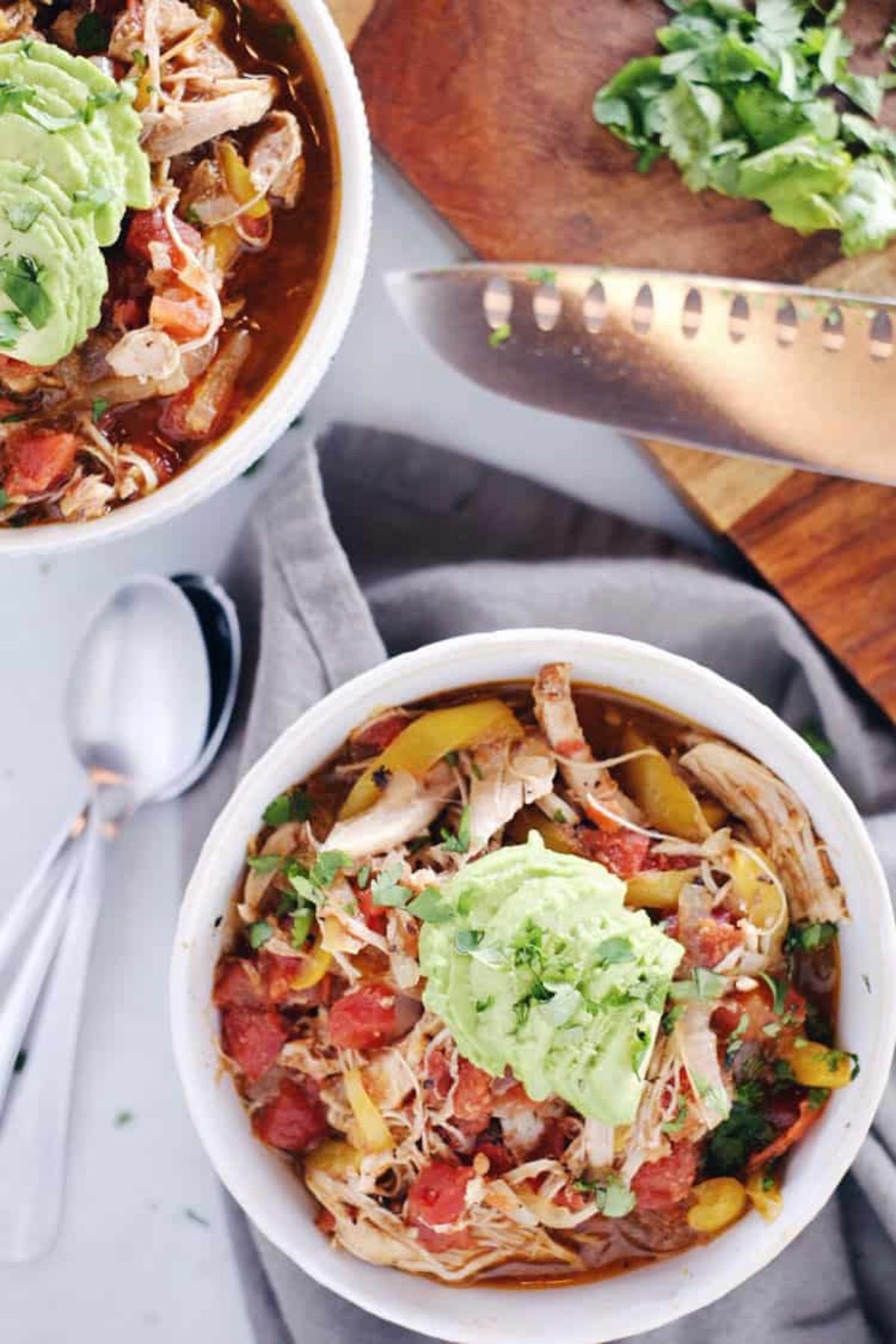 2 bowls of chicken soup with vegetables, topped with avocado and chopped herbs. silver spoons and a chopping knife sit on either side