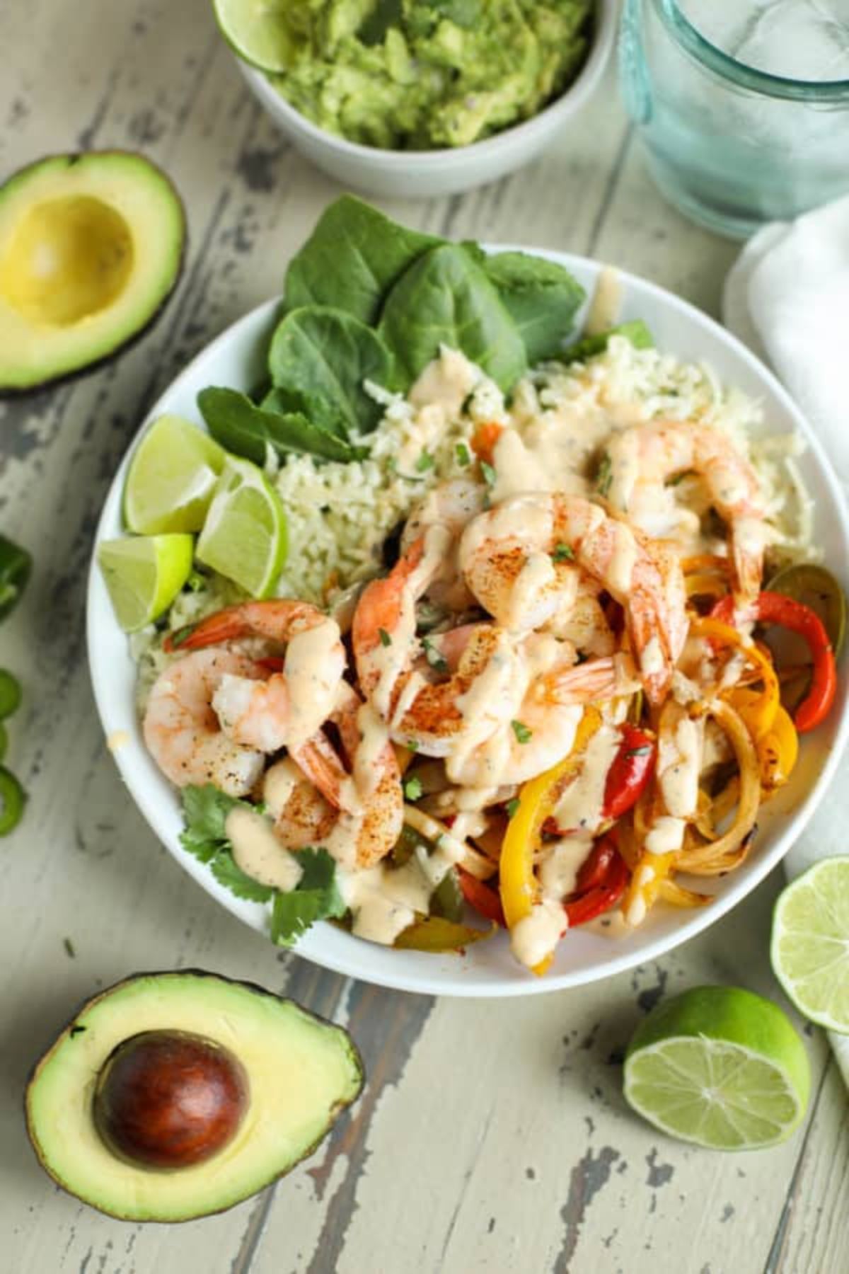 a bowl of rie with shrimp and sliced peppers on top, drizzled with reamy sauce and garnished with lime wedges. Halved avocados, lime wedges and a bowl of guacamole on the side