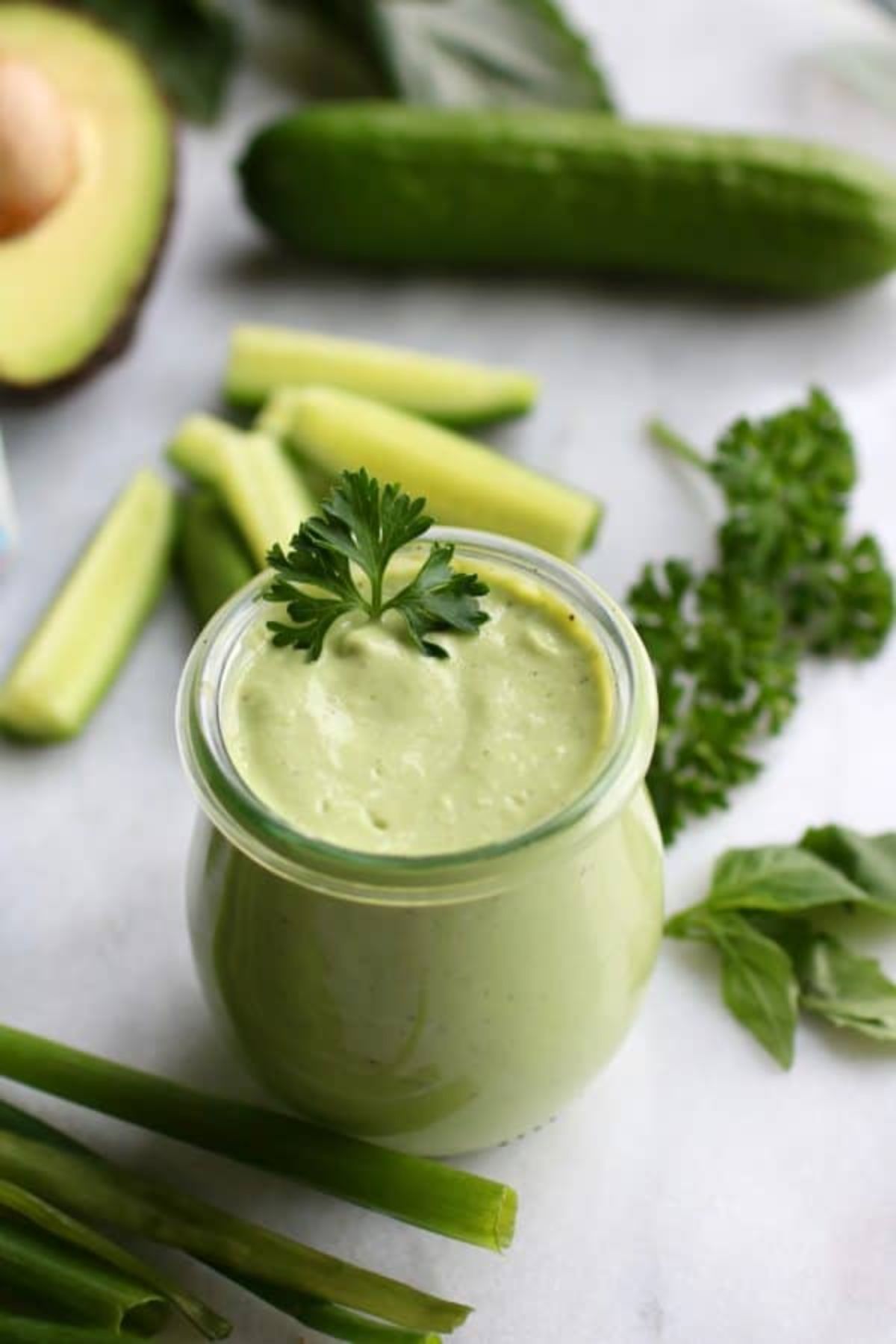 a glass jar of thick green dressing with a sprig of parsley in it. Behind is parsely, cucumber slices, a whole cucumber, scallions,a nd half an avocado