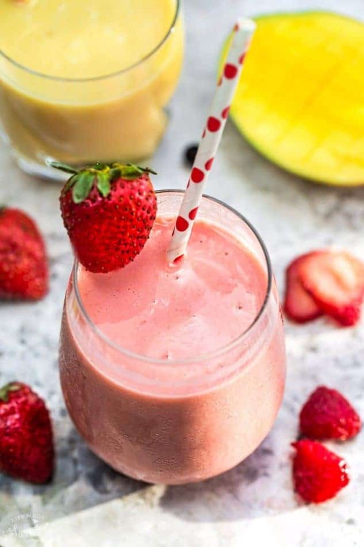 A glass of strawberry smoothie garnished with a strawberry and with a paper white and red straw in it. fresh strawberries are scattered around