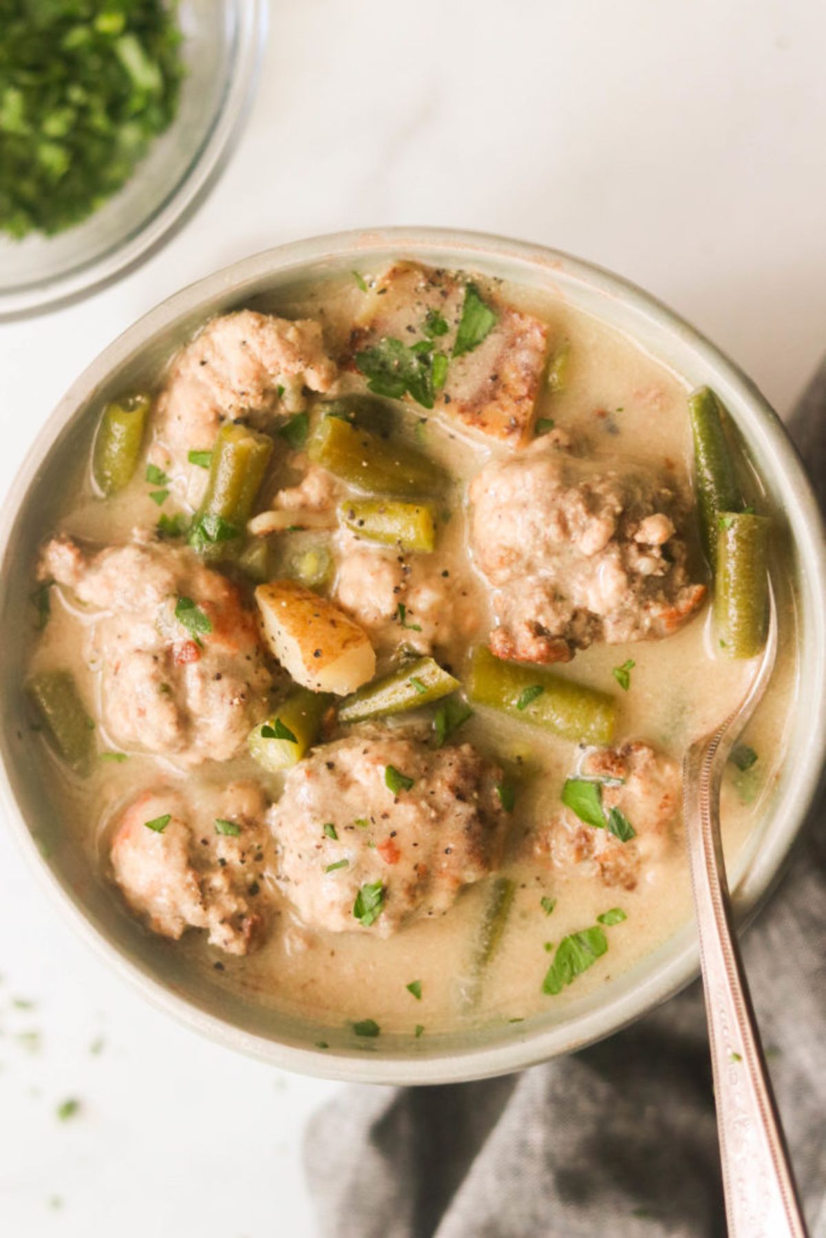 a white bowl of meatball creamy soup with chopped herbs. A spoon sits in the bowl