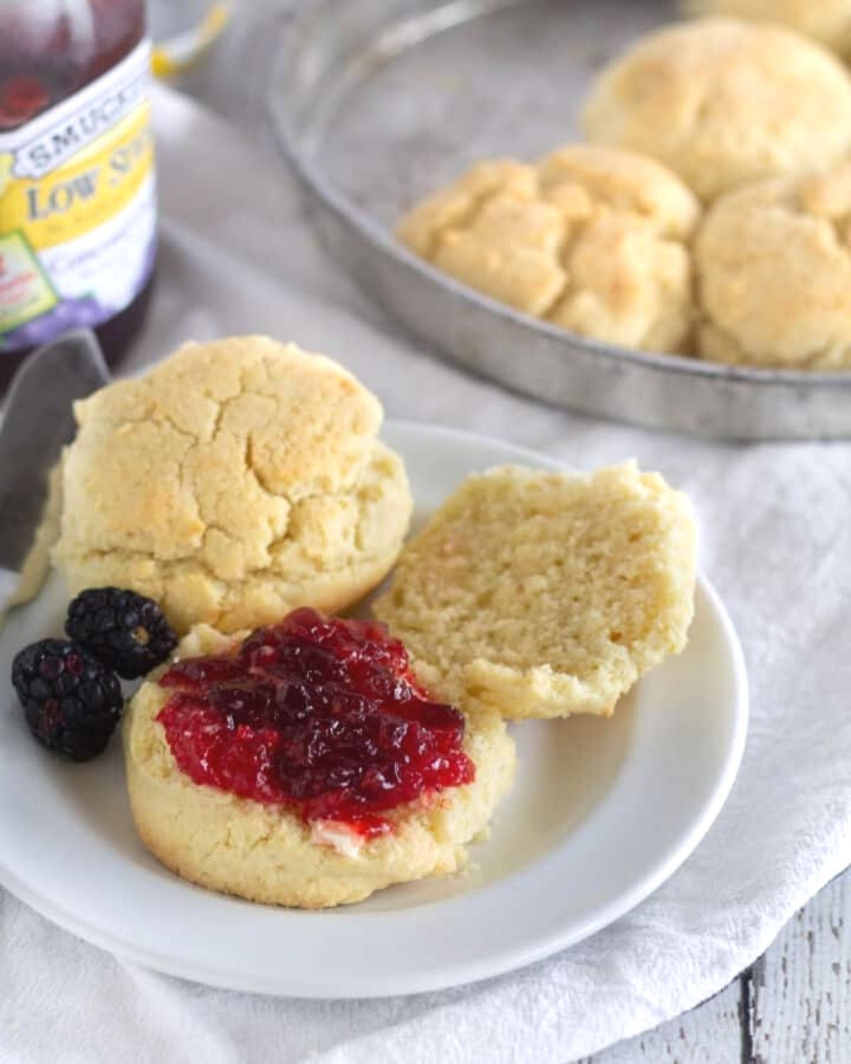 a plate with 2 almond flour biscuits, one cut in half and loaded with jam. In the background is a tin with more biscuts in it and to the left is a jar of low sugar jam