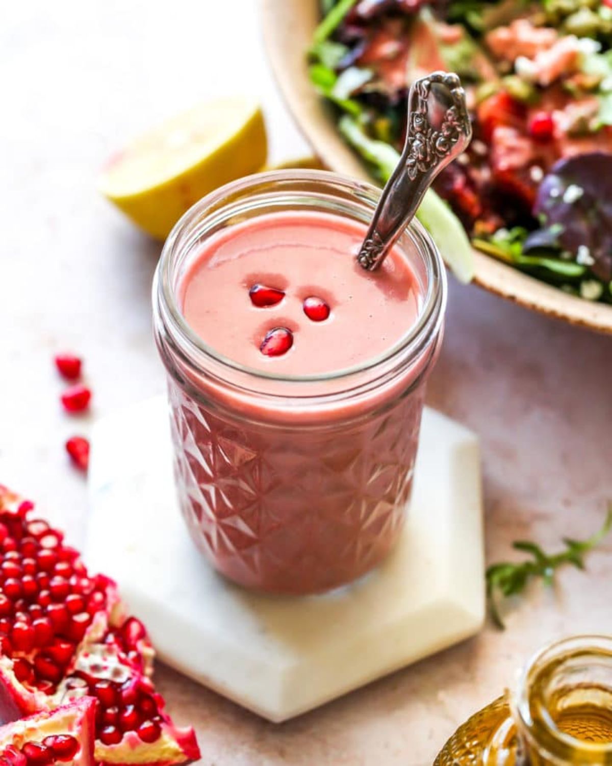 A glass jar of pink dressing with pomegranate seeds on the top and a silver spoon handle sticking out of it. Behind is a bowl of salad and a pomegranate