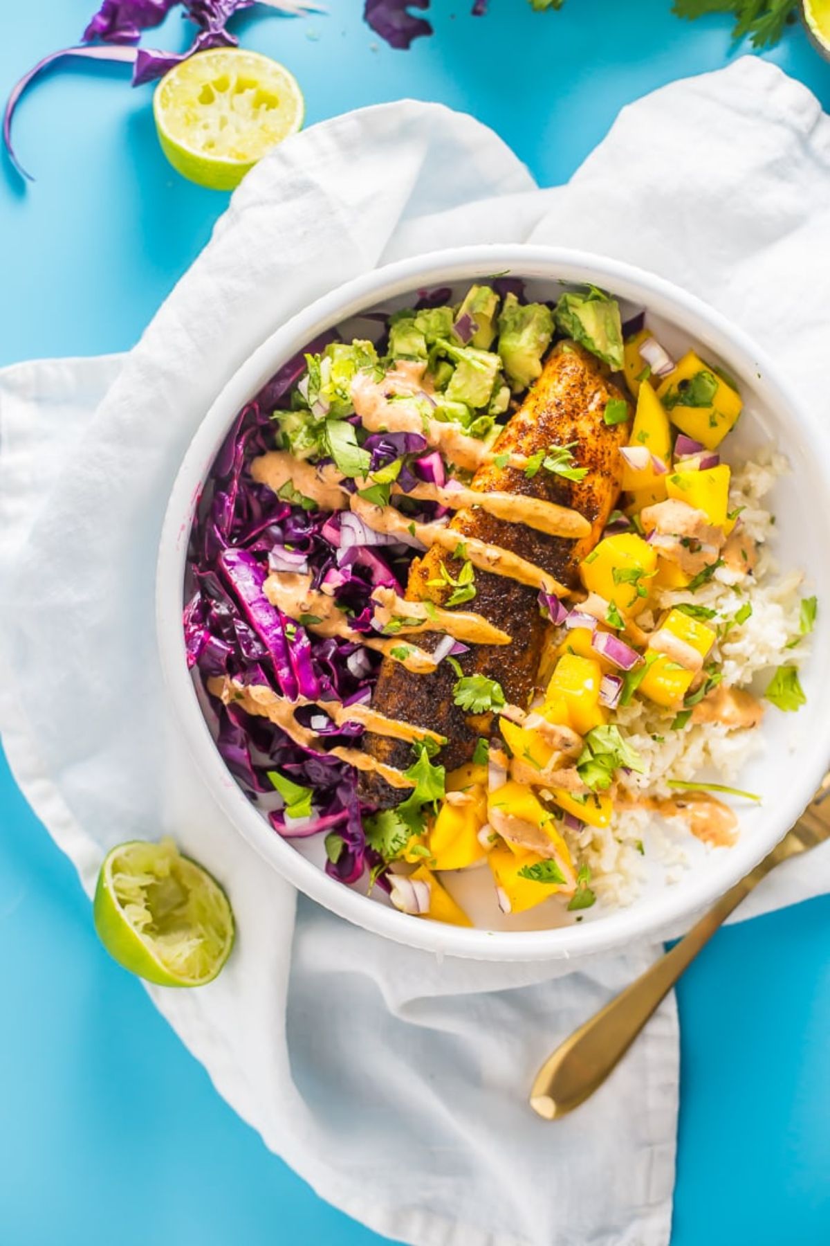 A whote bowl on a white cloth on a blue table. The bowl is filled with avocado, mango, red cabbage, a salmon filet, cauliflower rice and an orange sauce drizzled over everything