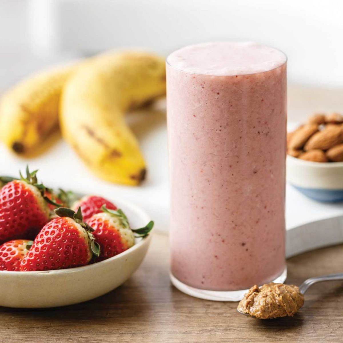 a glass of almond butter smoothie, next to a bowl of strawberries, 2 bananas and a bowl of almonds