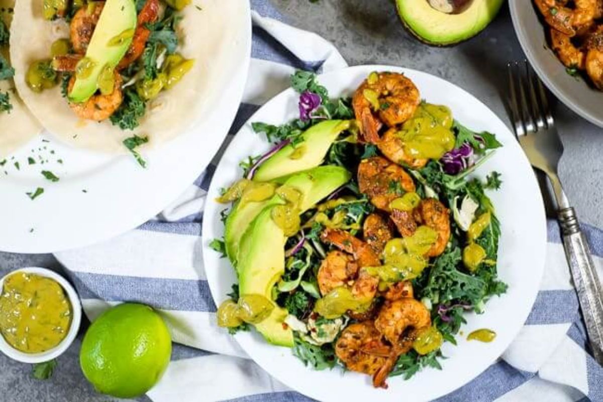 A white plate with kale, shrimp, avocado and lime sauce srizzled over it. THe plate is on a blue and white striped cloth