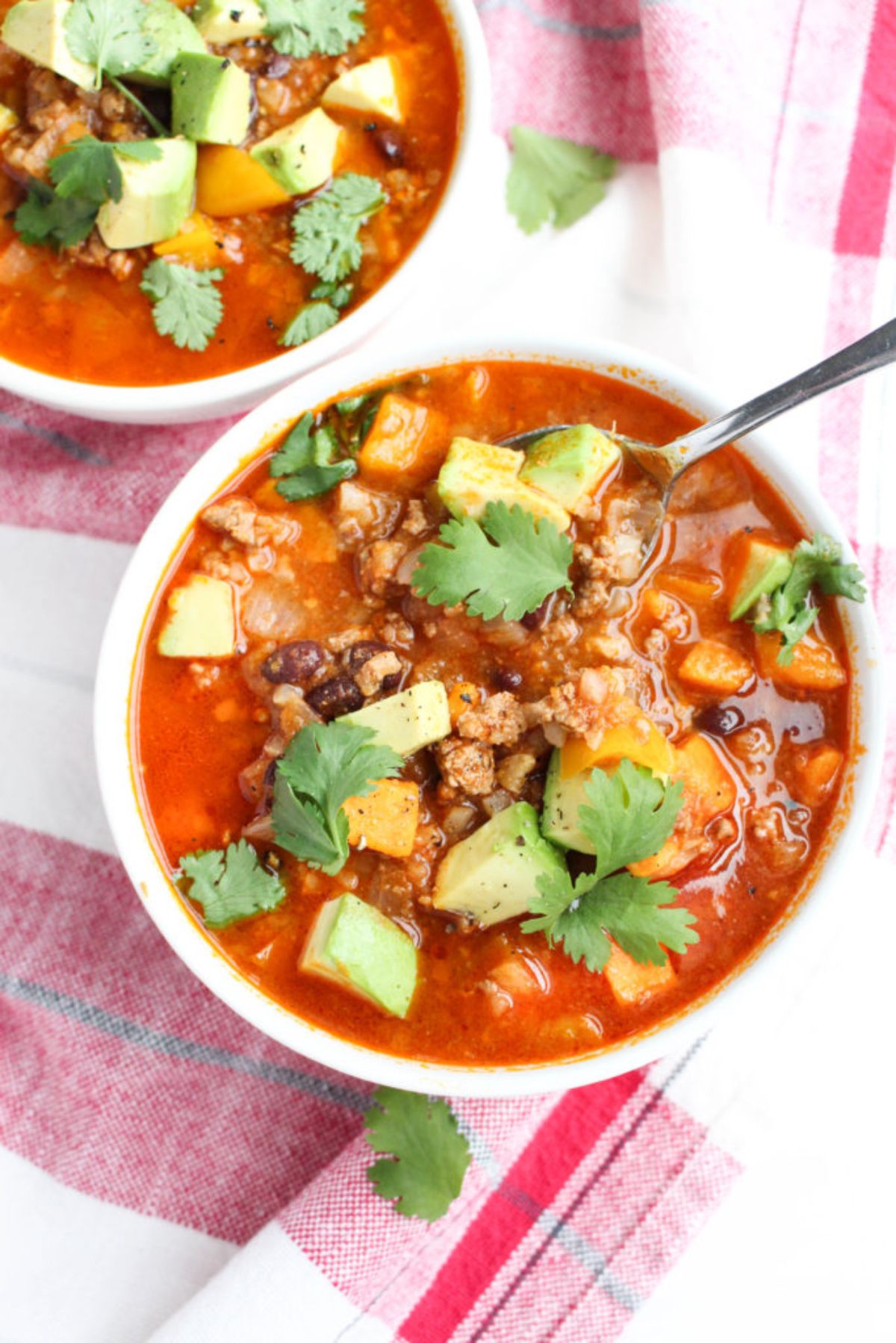 2 white bowls of taco soup toppped with avocado and herbs, a silver spoon resting in one bowl