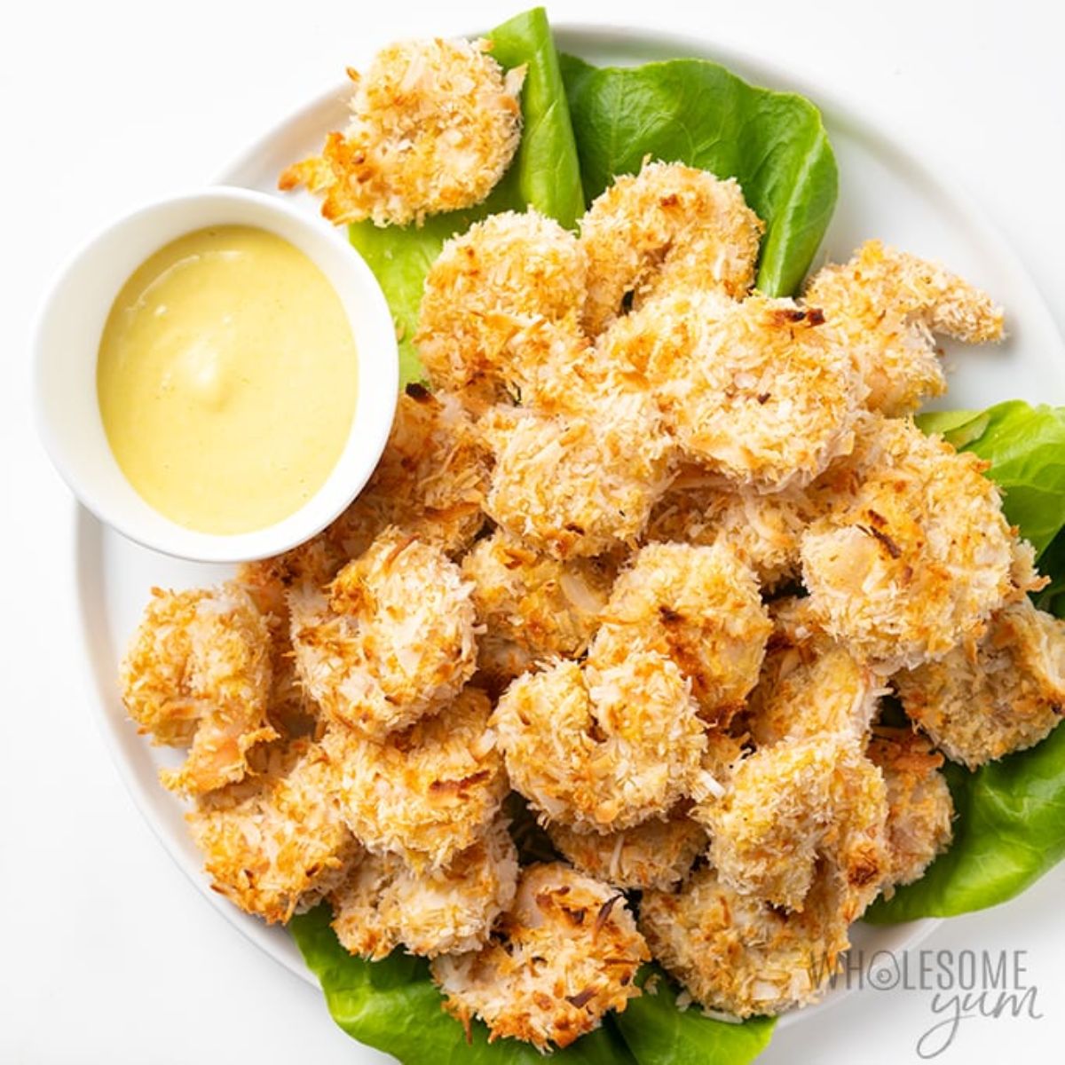 A plate of coconut shrimp on a bed of lettuce leaves with a white pot of dip to the side