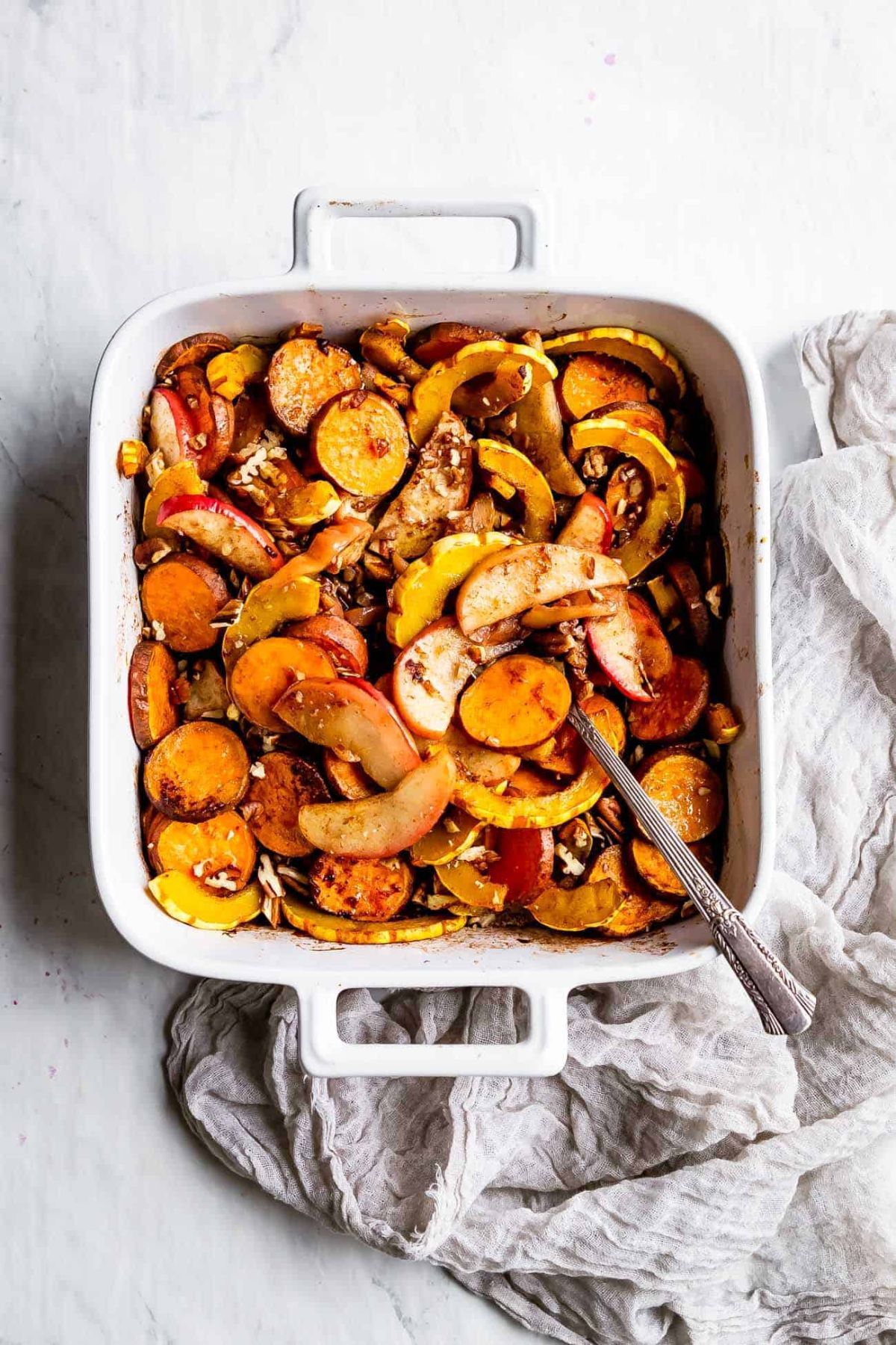 a square oven dish with slices of baked squash, sweet potato and apple in it. A silver spoon is sticking out of the dish