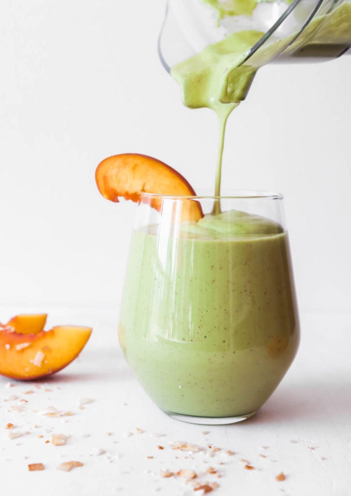 A glass with green smoothie being poured into it from a blender jug, the glass is garnished by a peach slice