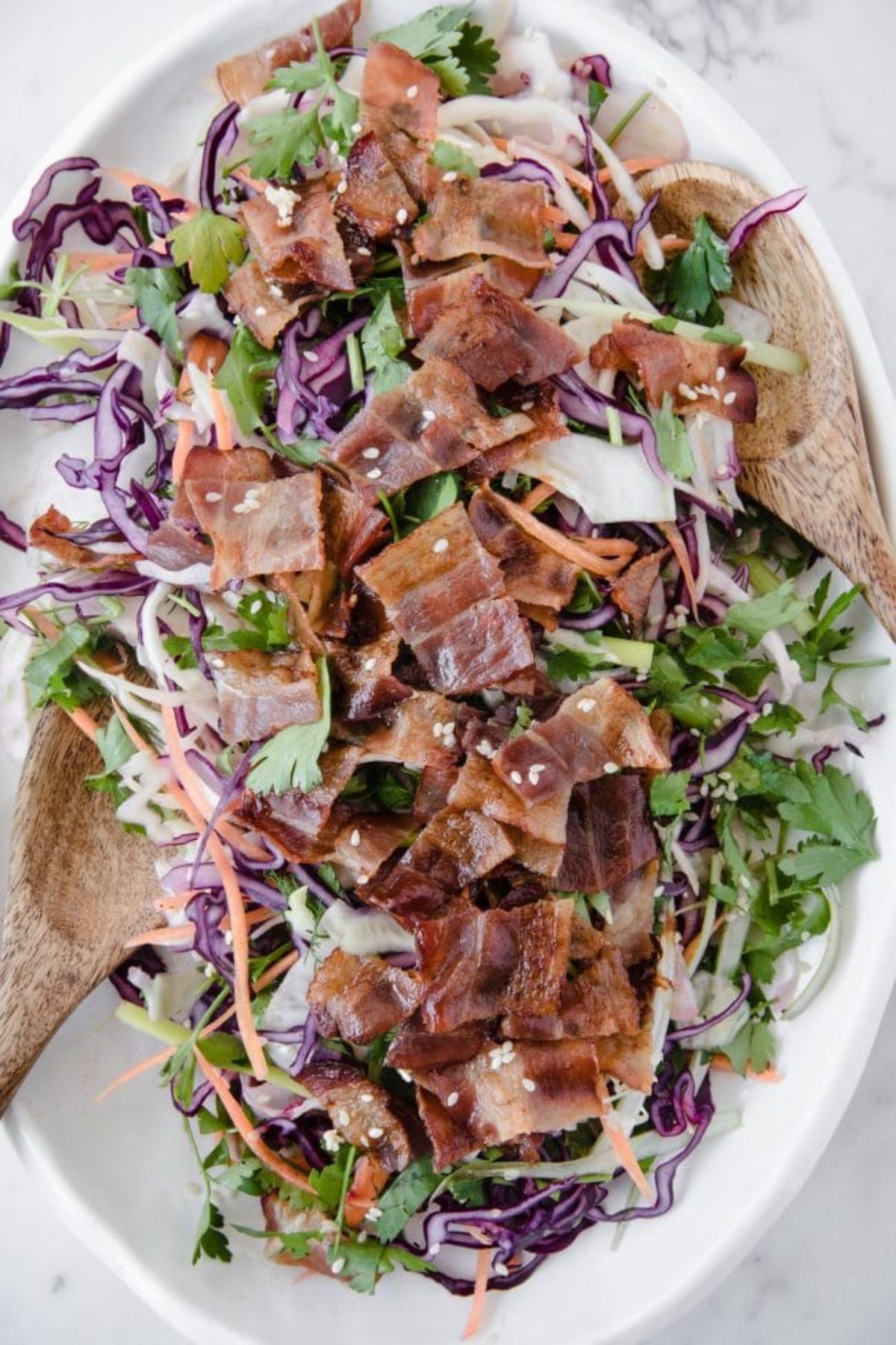 An oval white dish with bacon and salad. A wooden spoon is resting on both sides