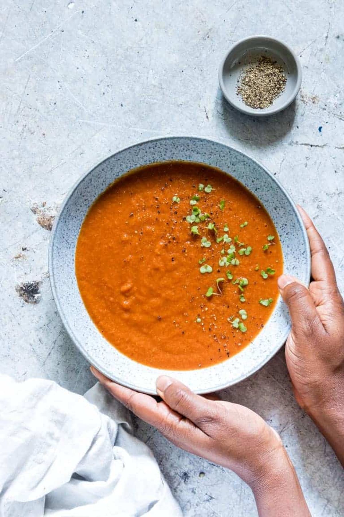 a light blue bowl held by two hands. Inside is tomato soup with herbs sprinkled on top.