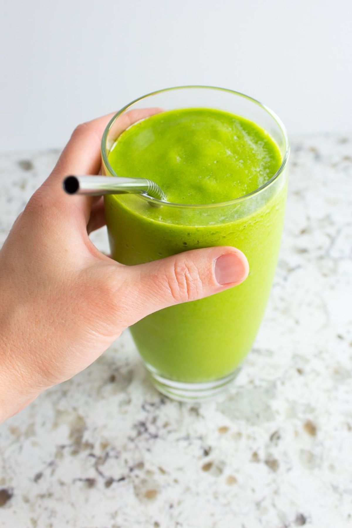 A hand holds a glass of green smoothie with a metal straw sticking out of it