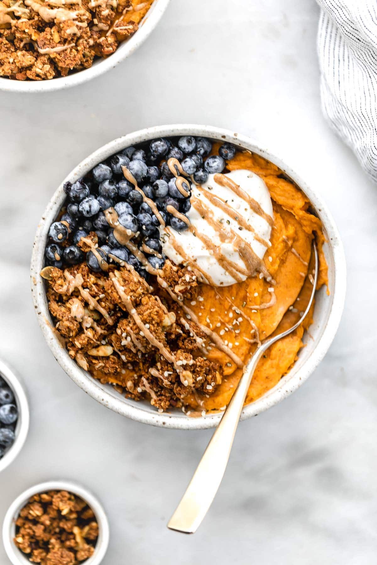 a bowl of sweet potato puree, blueberries, granola and yoghurt, drizzled with sauce and with a spoon on the side