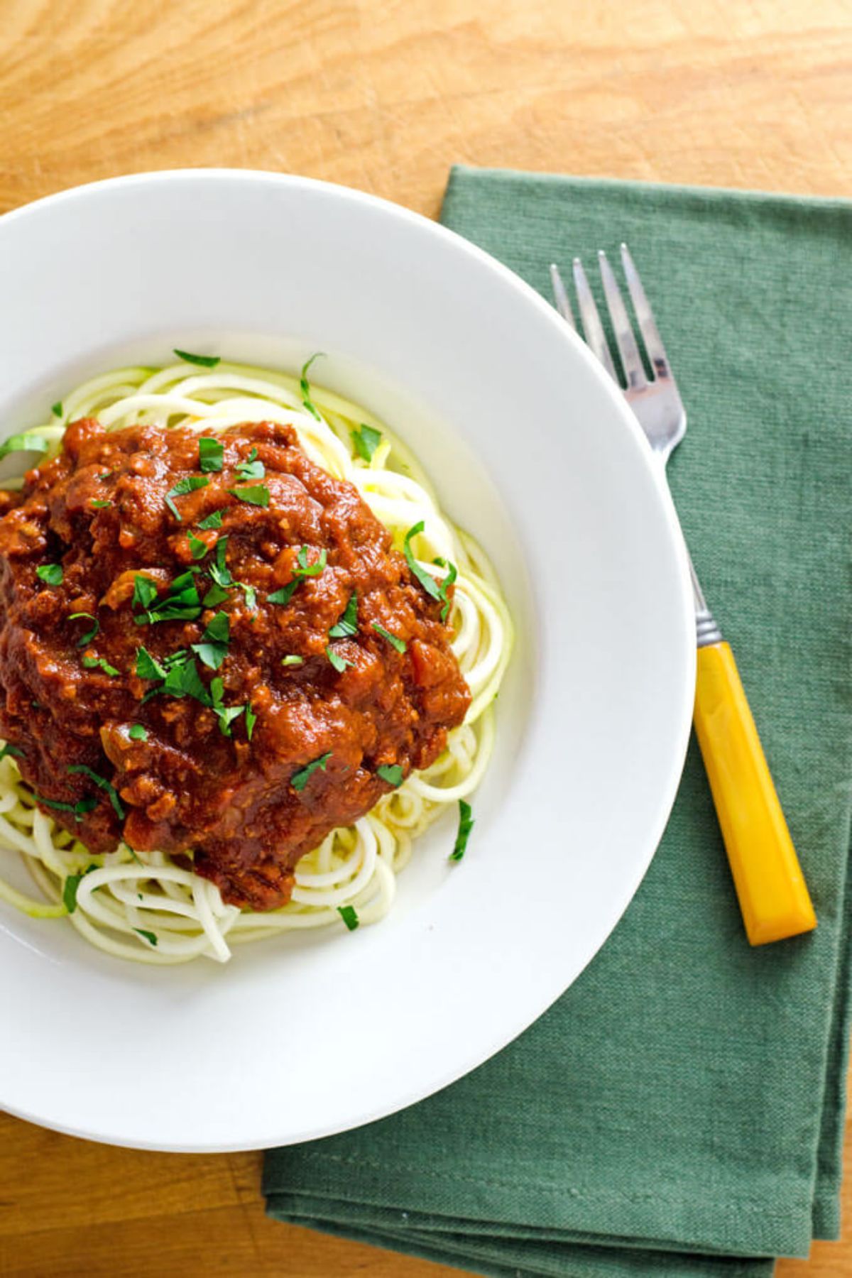 A white plate resting on a green cloth next to a yellow handled spoon. The plate contains bologense sauce sitting on a pile of zucchini noodles