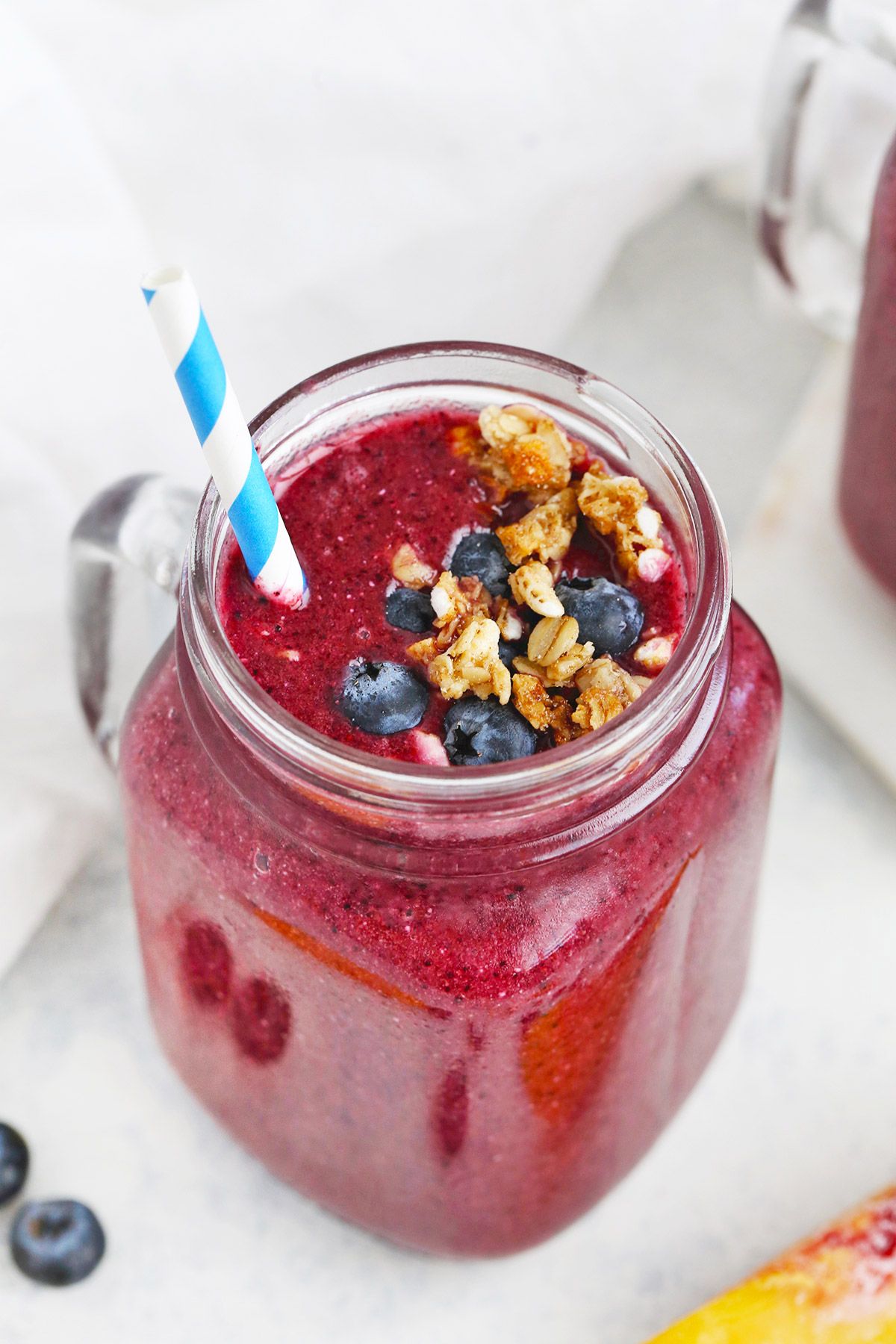 A squared mason jar filled with blueberry peach smoothie, topped with granola and fresh bluberries, with a blue and white striped straw sticking out