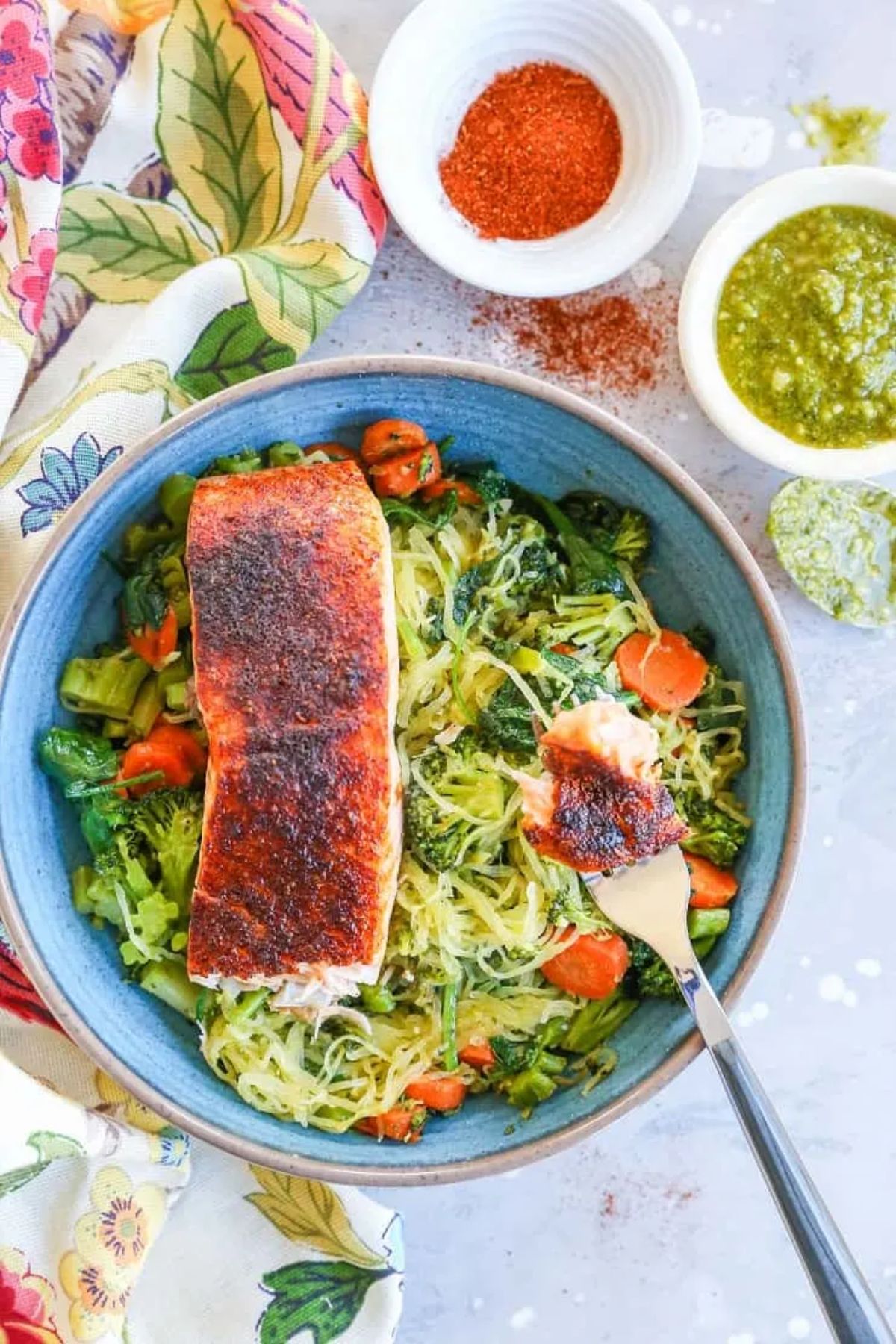 a blue bowl od spaghett squash noodles, with chopped vegetables and a fillet of salmon on top. 2 white bowls of spices and sauces are to the side