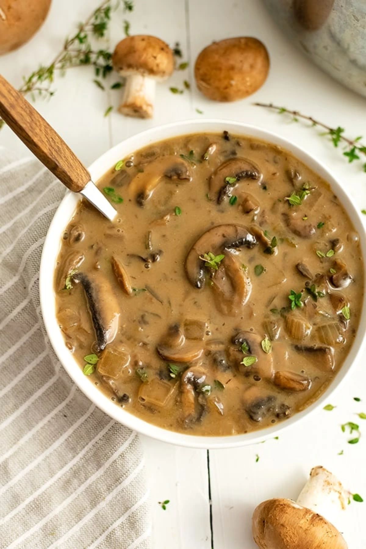 a white bowl of chunky mushroom soup and a wooden-handled spoon resting in it. Th ebowl is surrounded by mushrooms and rosemary sprigs