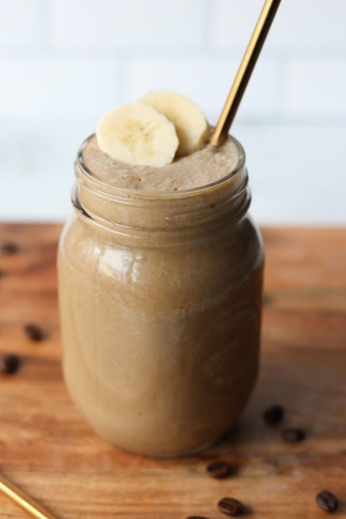 A mason jar filled with banana coffee smoothie, topped with banana slices and a gold metal straw sticking out