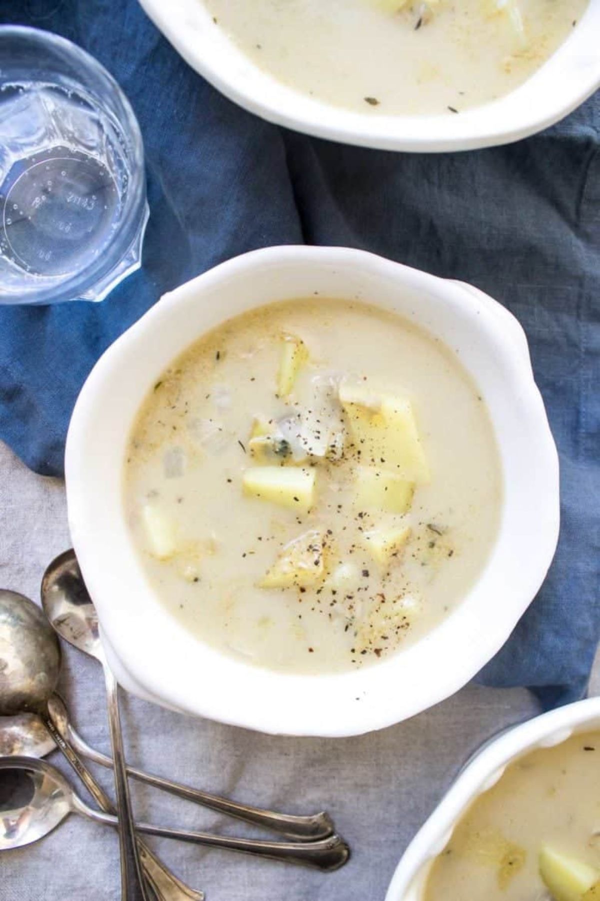 a white bowl of clam chowder sits on a blue cloth with a pile of spoons next to it