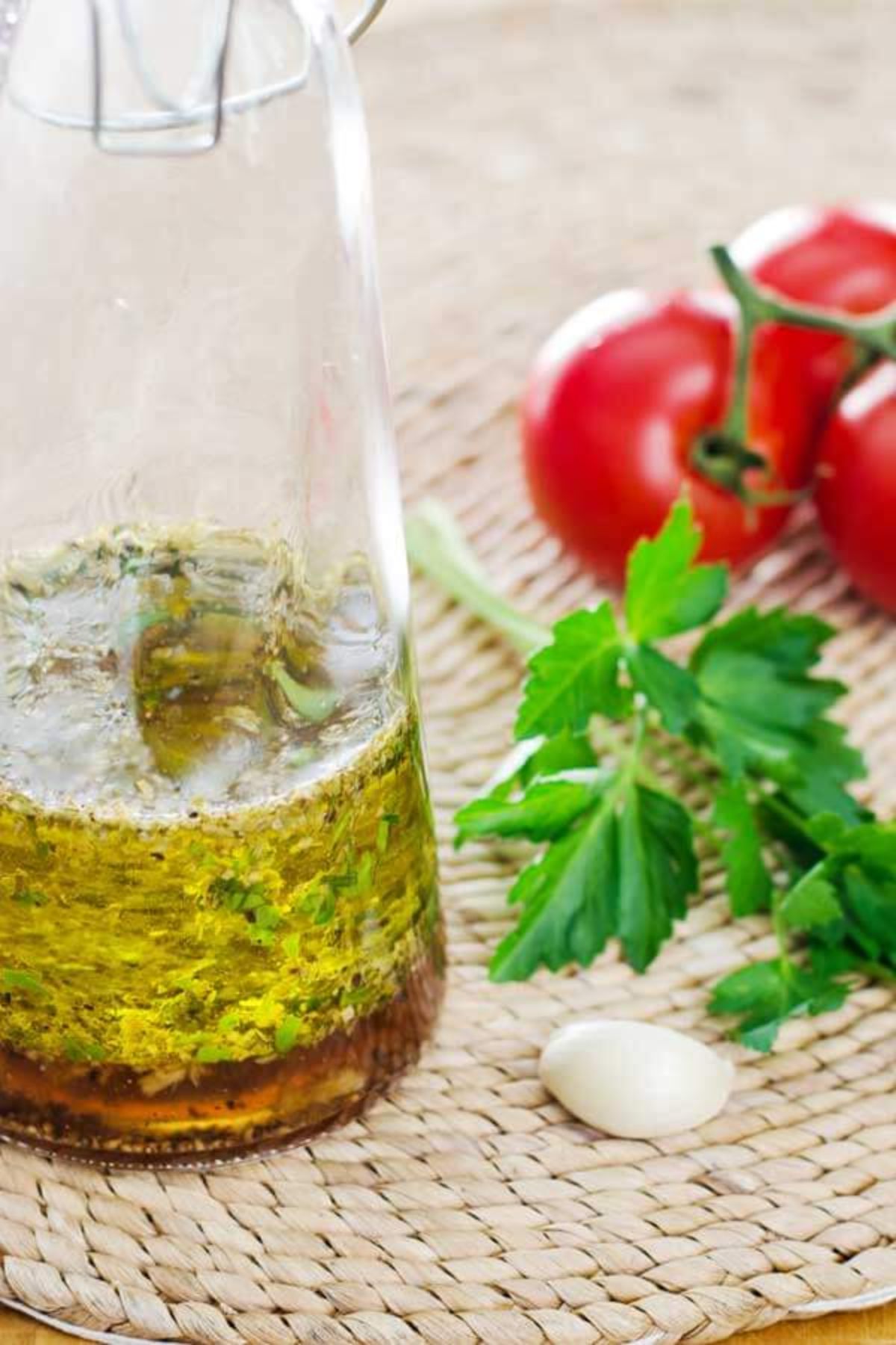 A tall glass flask of vinaigrette with a parlsey sprig, a garlic clove, and a bunch of tomatoes next to it on a woven mat