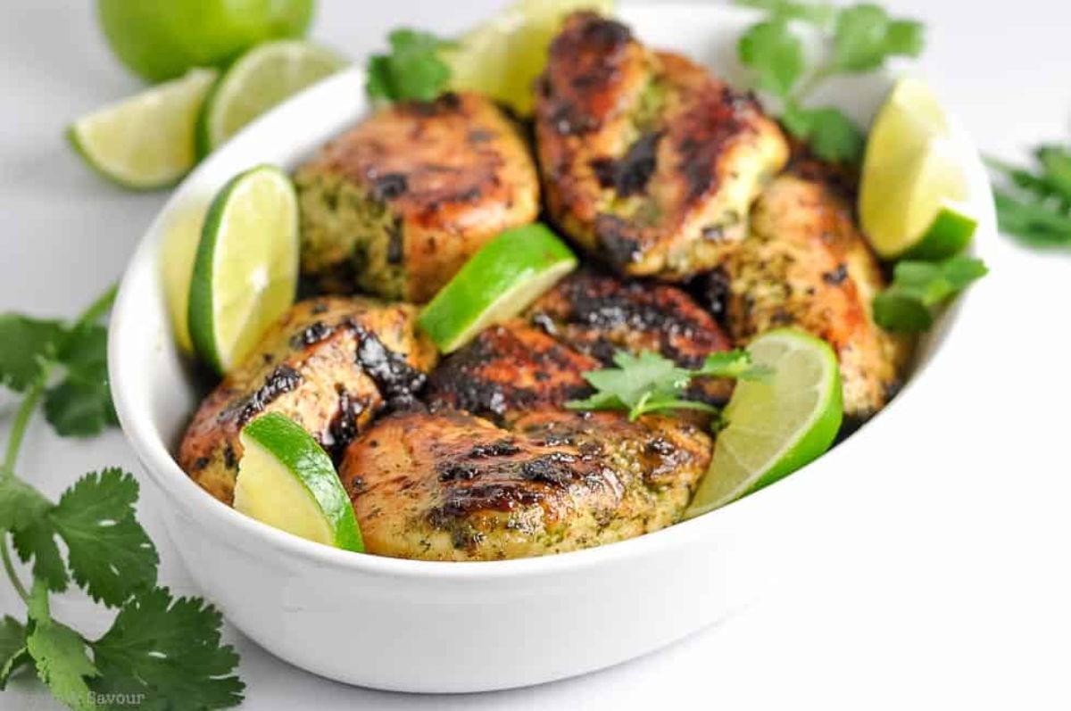 a white casserole dish full of roasted chicken thighs, garnished with chopped herbs and lime slices