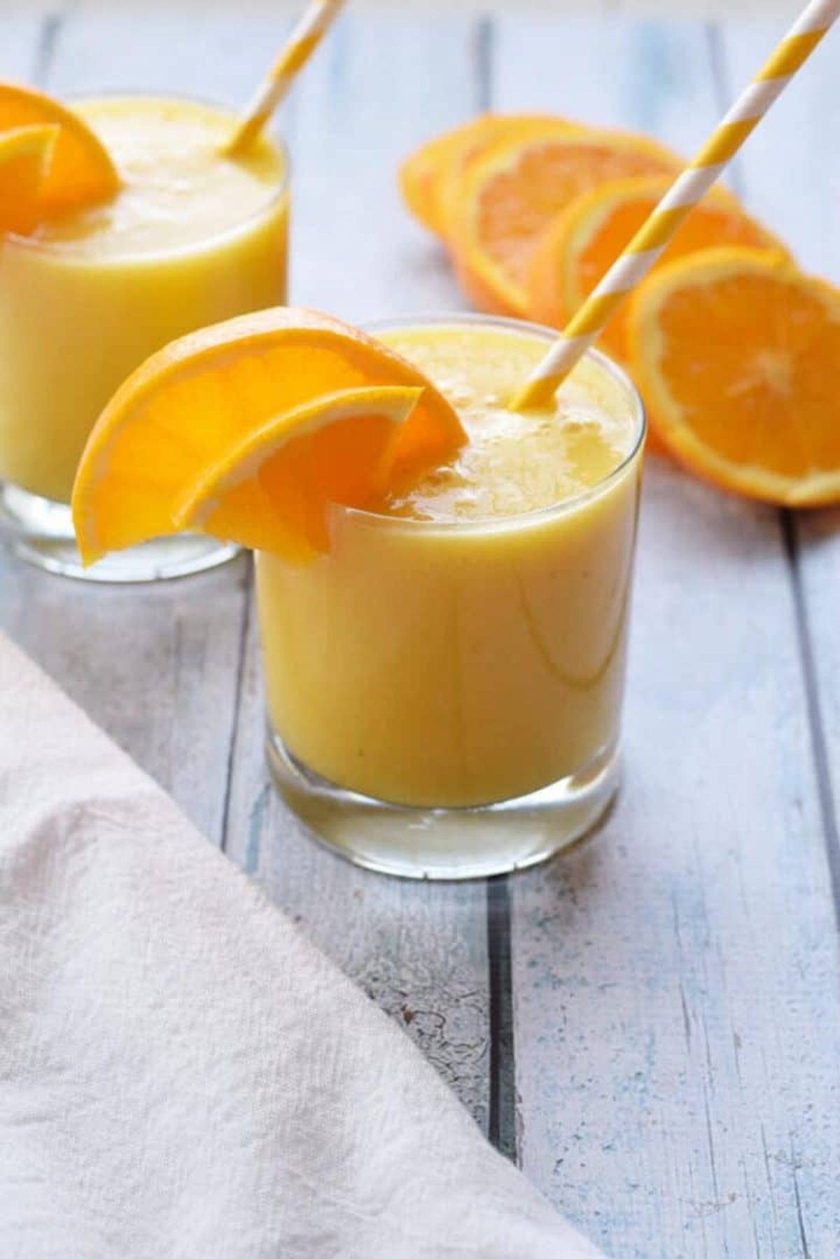 2 glasses of orange smoothie, garnished with orange slices and with orange and white striped straws sticking out. Oranges slices can be seen in the background