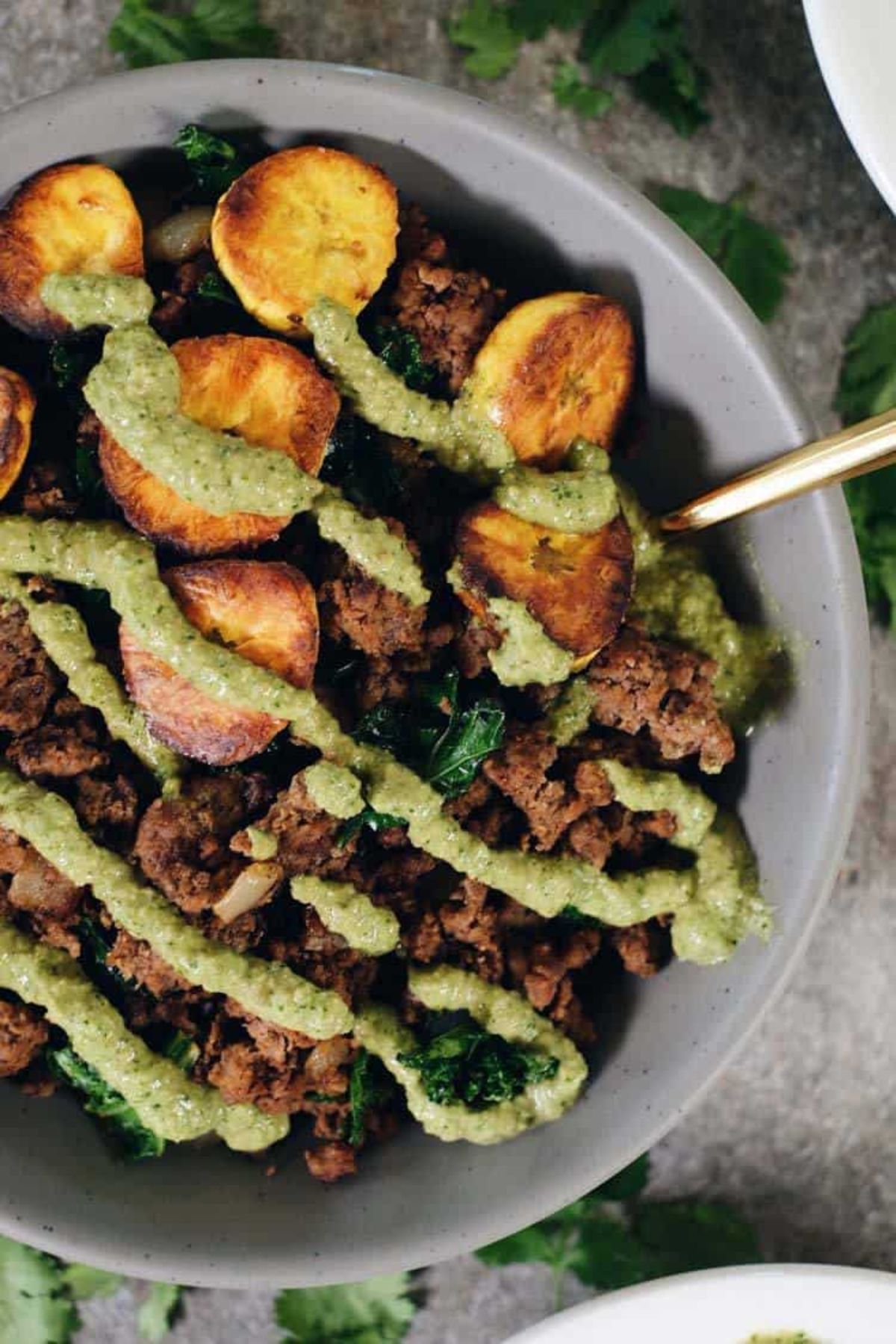 A grey bowl of beef and plantain casserole drizzled with green pesto