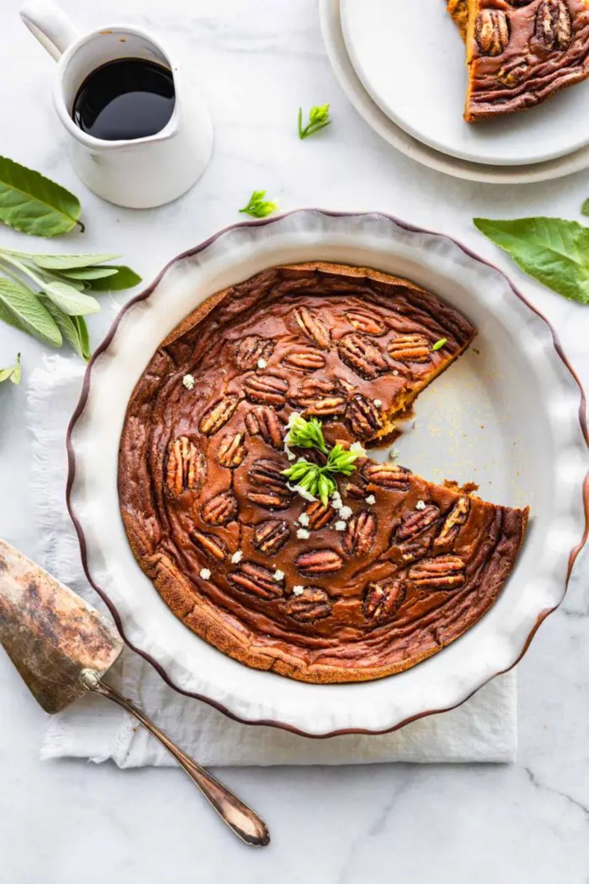 a pie dish with crustless sweet potato pie in it, next to a cake slice and a jug of sauce. Sage leaves are scattered around