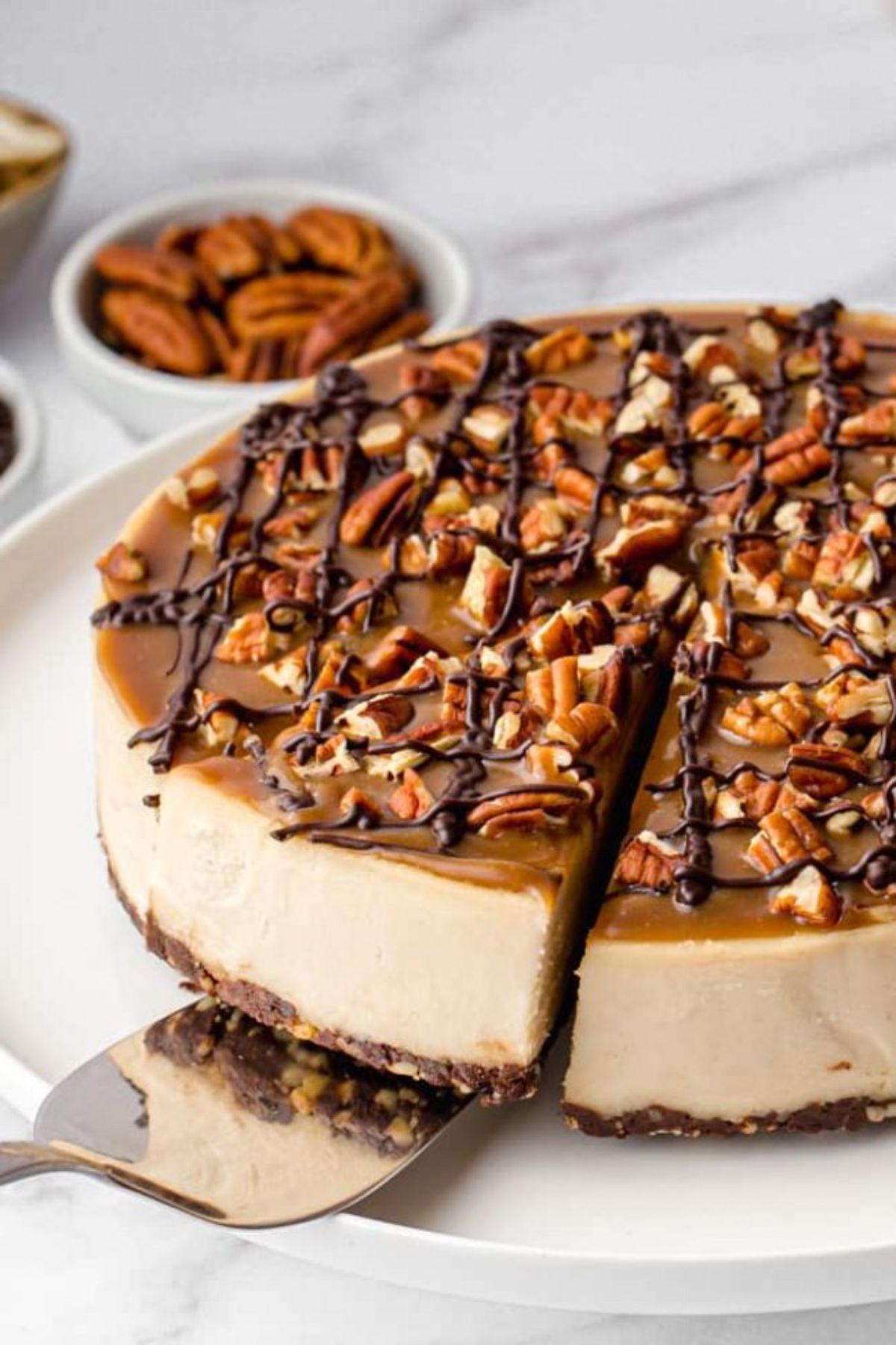 A plate of caramel coloured cheesecake with one segment cut out and being liften out by a cake slice. A bow of ppecans is in the back