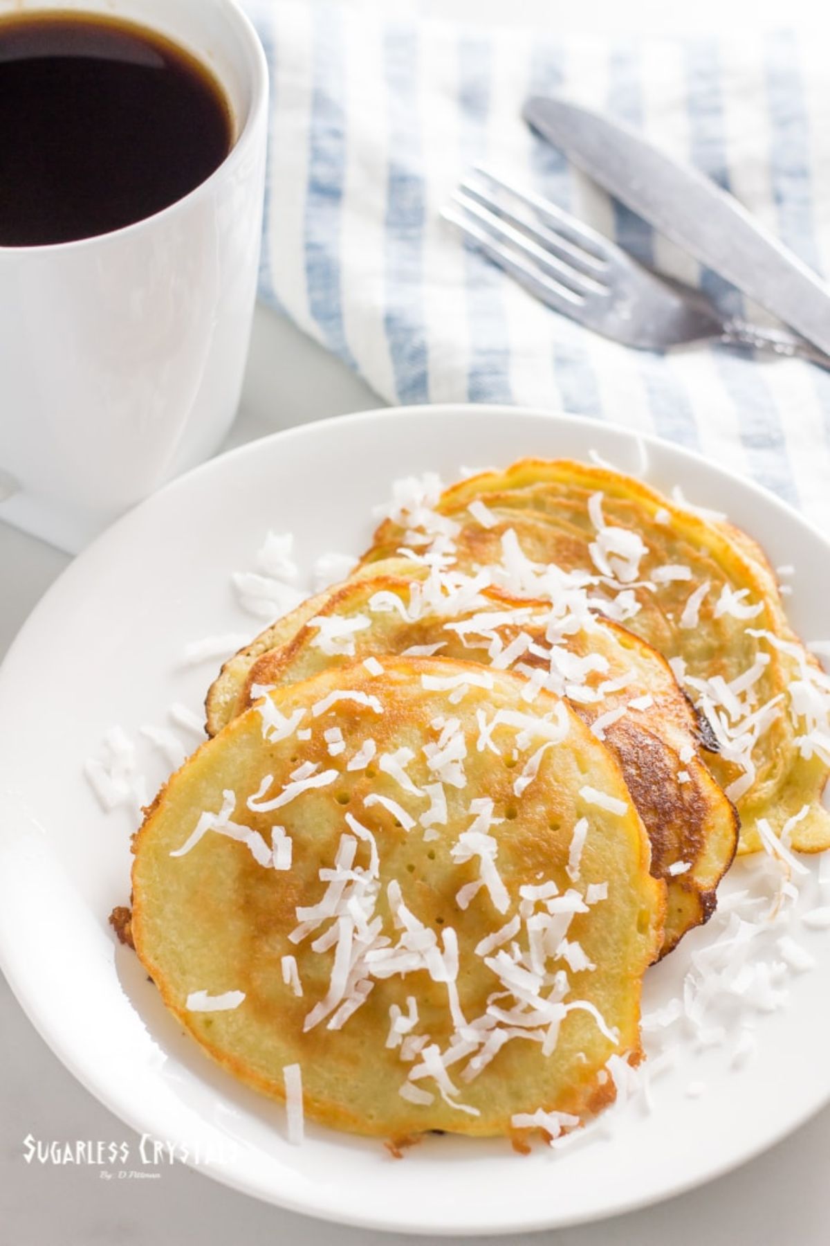 A white palte with pancakes on it, sprinkled with shreded coconut. A cup of coffee and a knife and fork are behind it