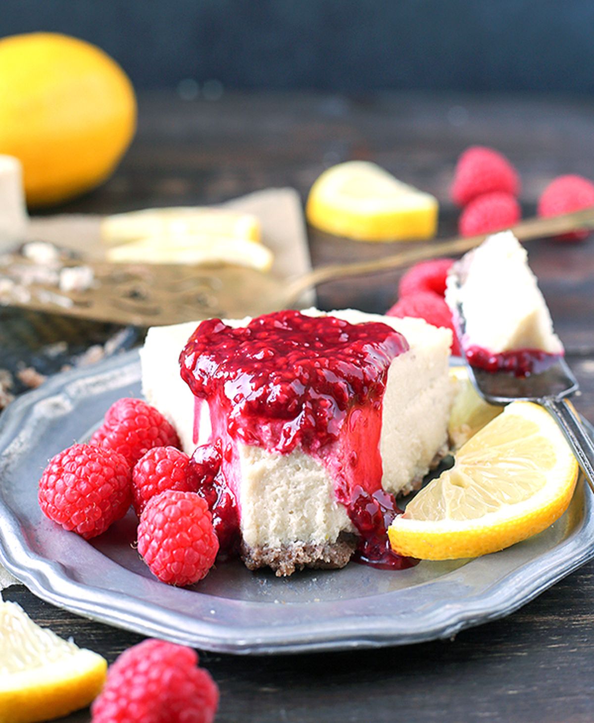 A blue plate with a segment of vanilla cheesecake drizzled with raspberry coulis and surrounded by lemon slices and fresh raspberries