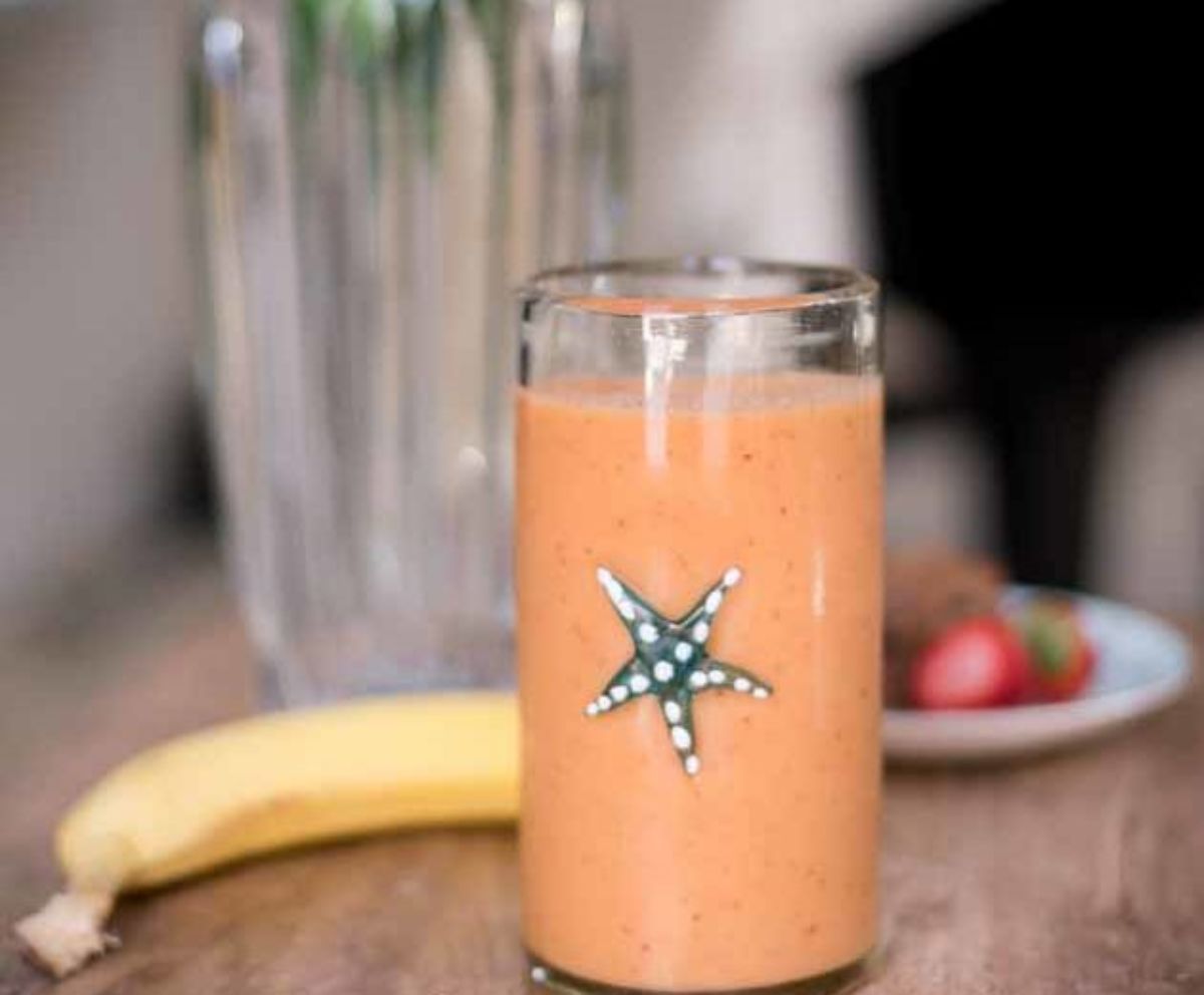 A glass of carrot apple and banana smoothie A banan and some frsh straberries can be seen behind