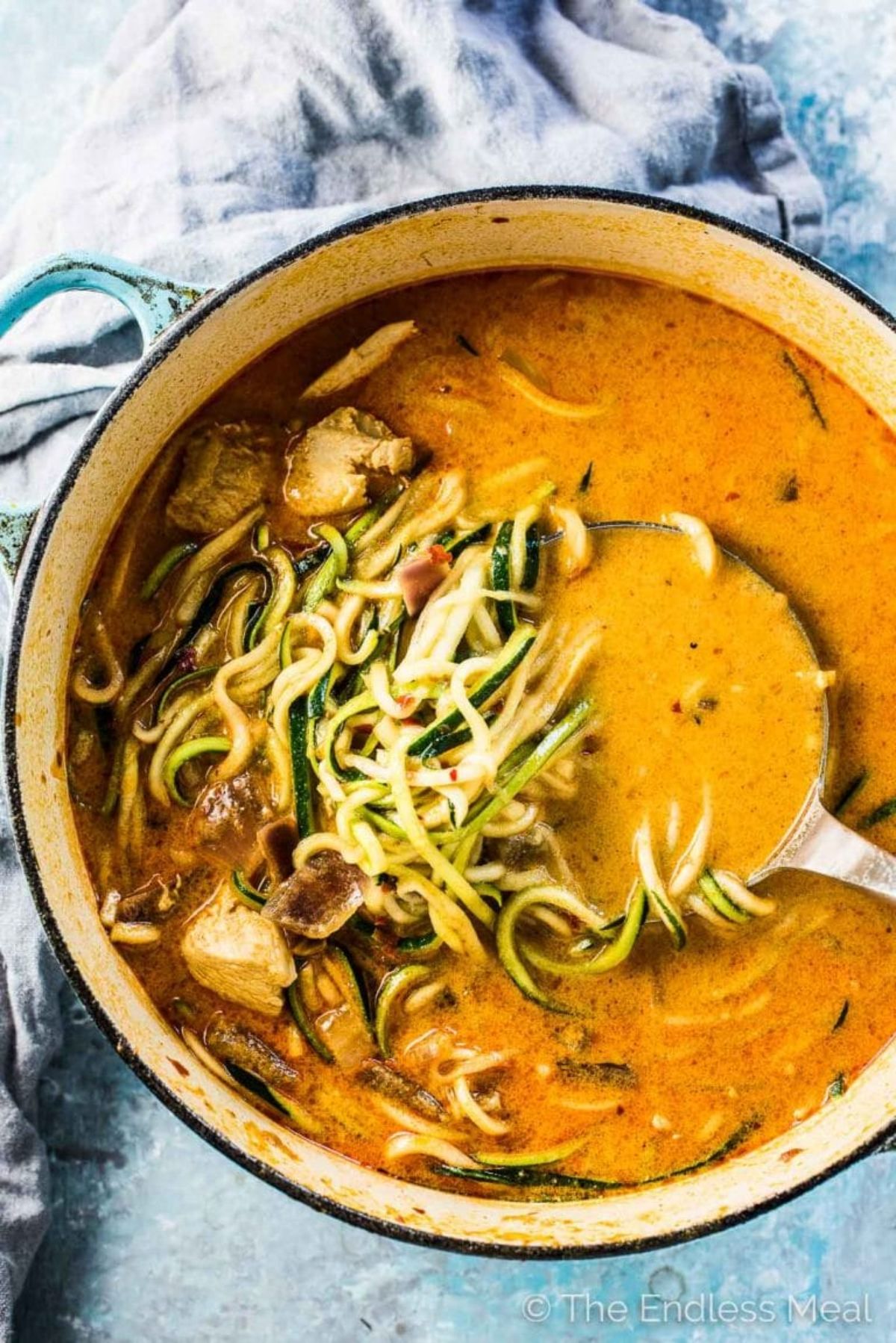 a pot of red soup with chicken andzucchini noodles seen. A ladel is resting in the pot