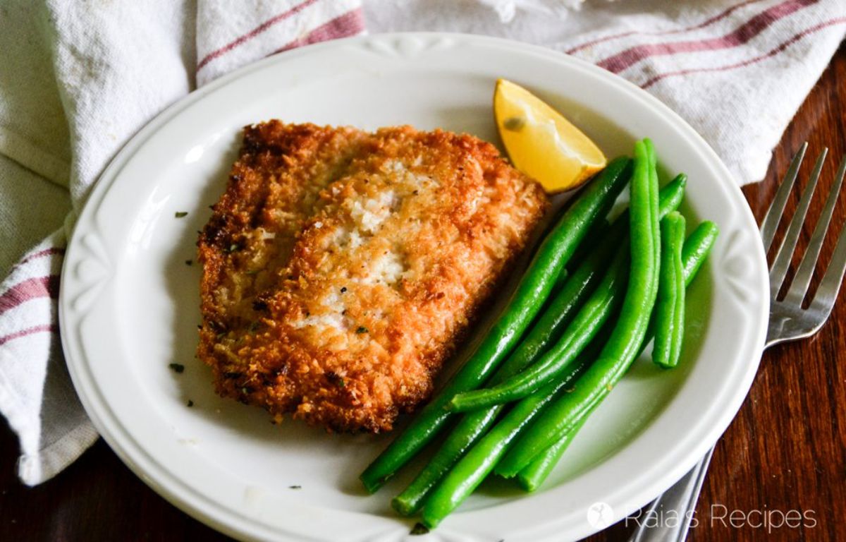 a white plate with a breaded fish fillet and green beans, garnished with a lemon edge